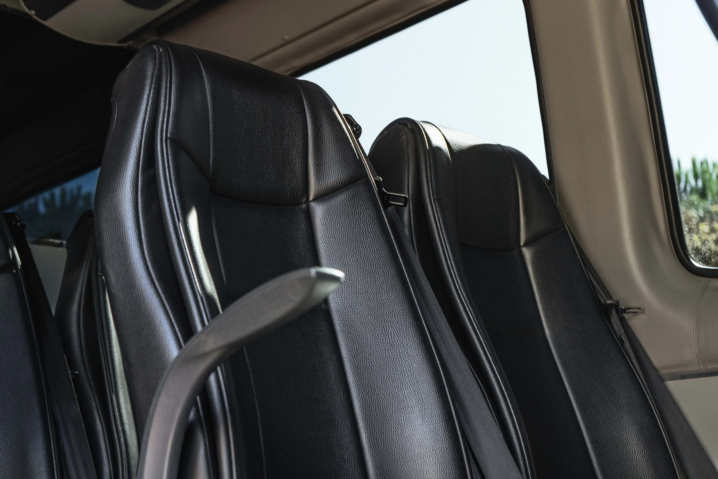 Assentos de couro preto na parte interna de um ônibus ou van, com janelas ao lado.