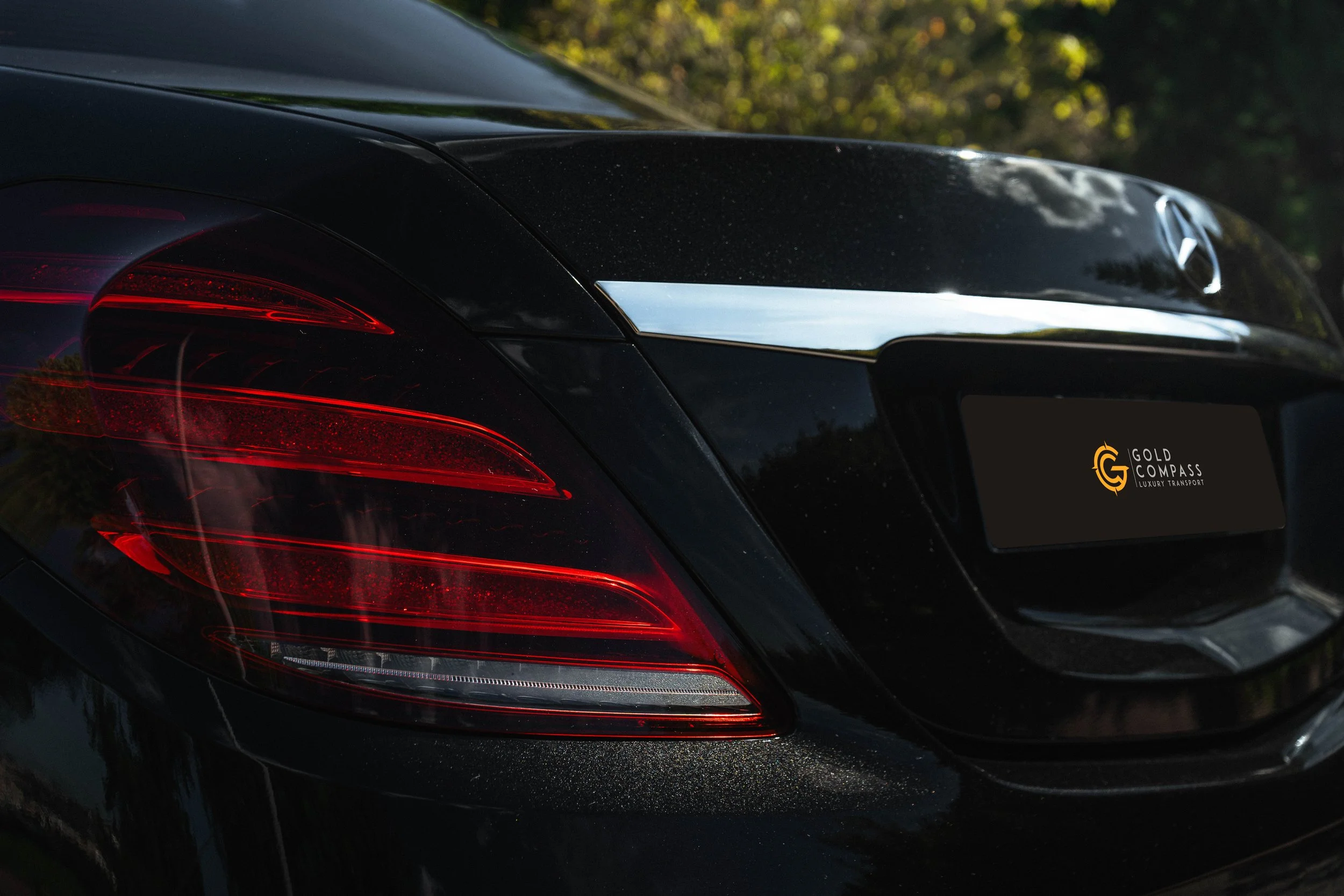 Parte traseira de um carro preto de luxo, mostrando a lanterna vermelha e a área do porta-malas com uma placa que diz 'Gold Compass Luxury Transport'.