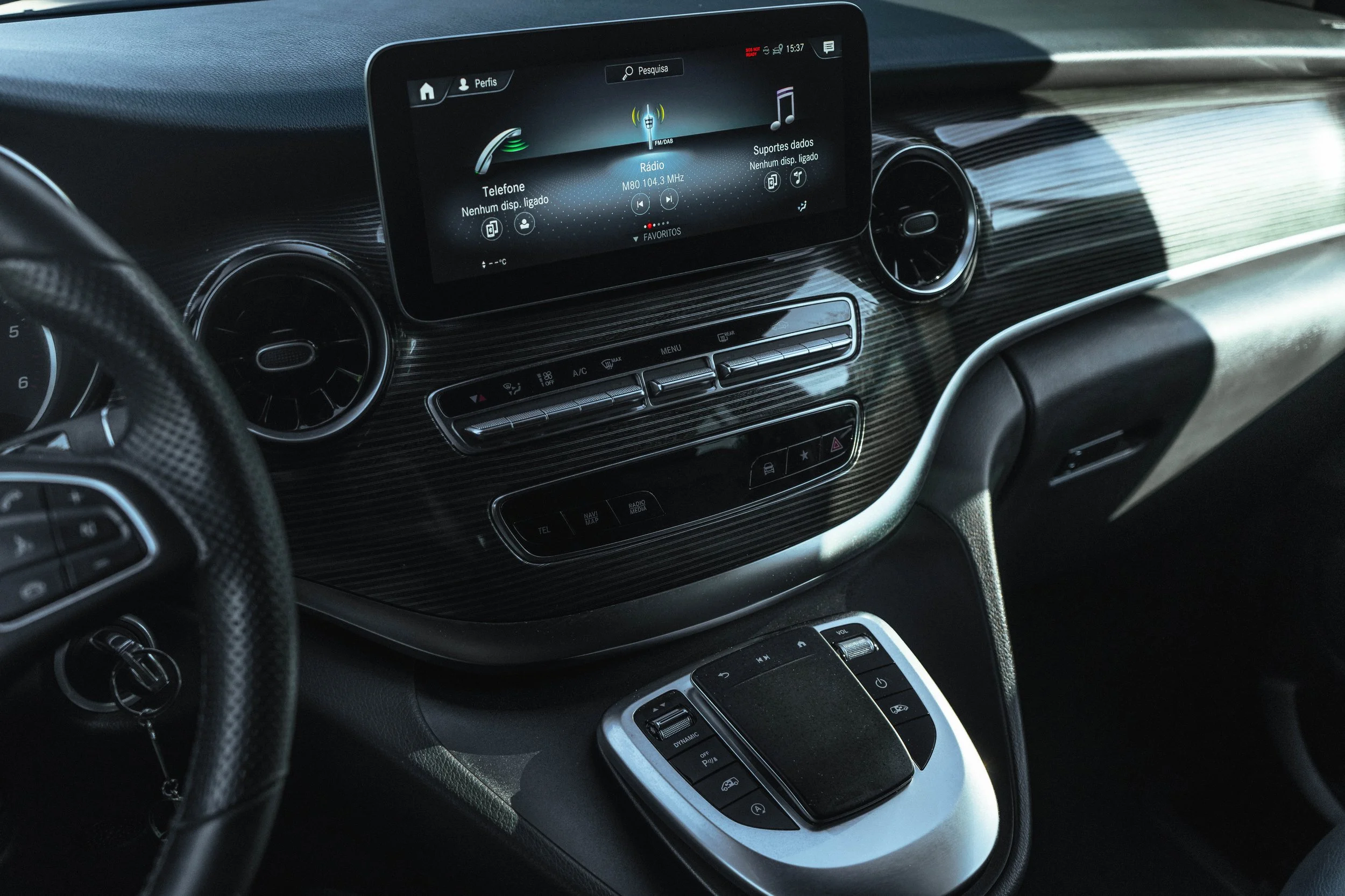 Interior de um carro, mostrando o painel central com tela digital, controles de cruzeiro e volante, detalhes em madeira e metal