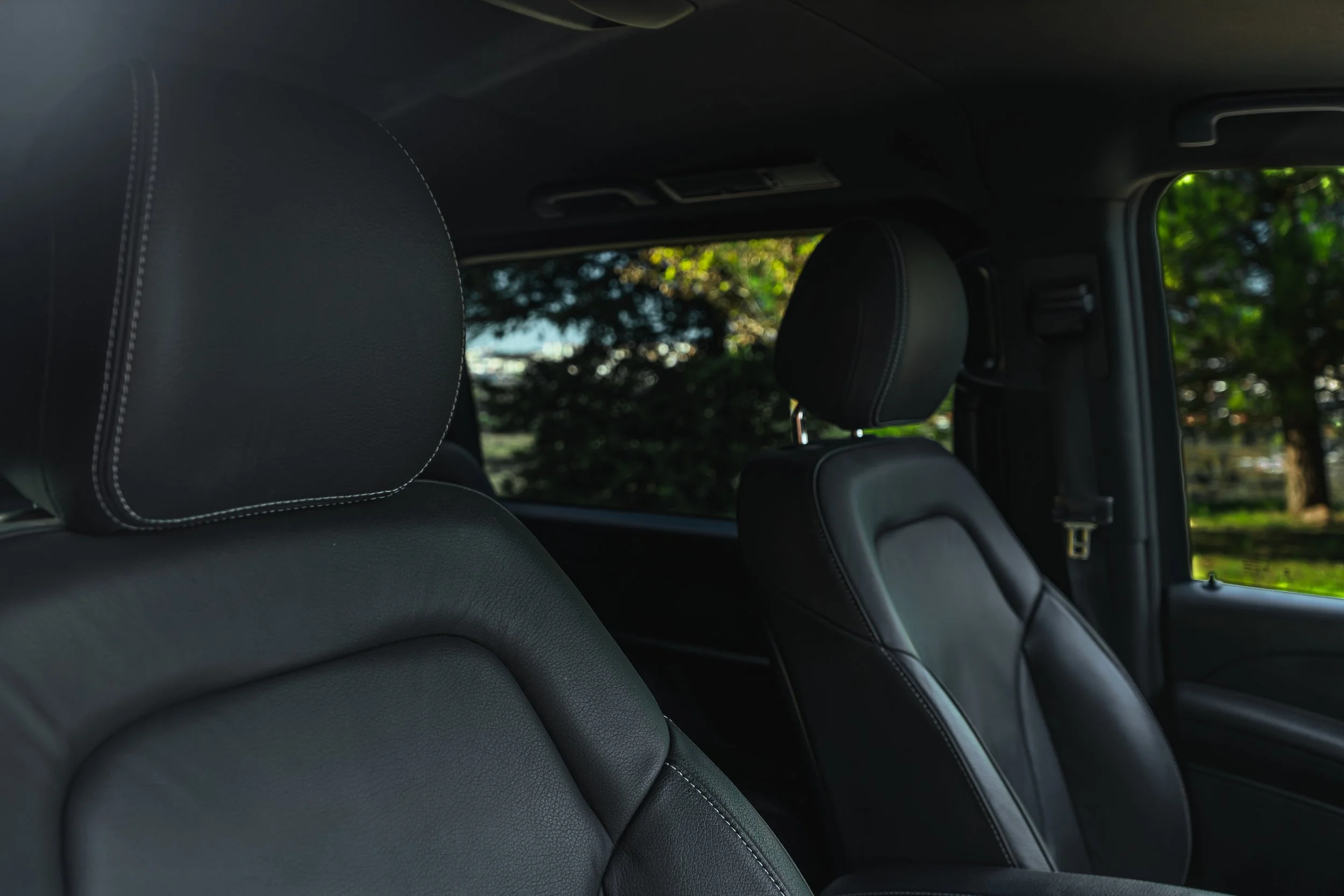 Interior de um carro com assentos de couro preto e cabeça de apoio, com janela mostrando árvores ao fundo.