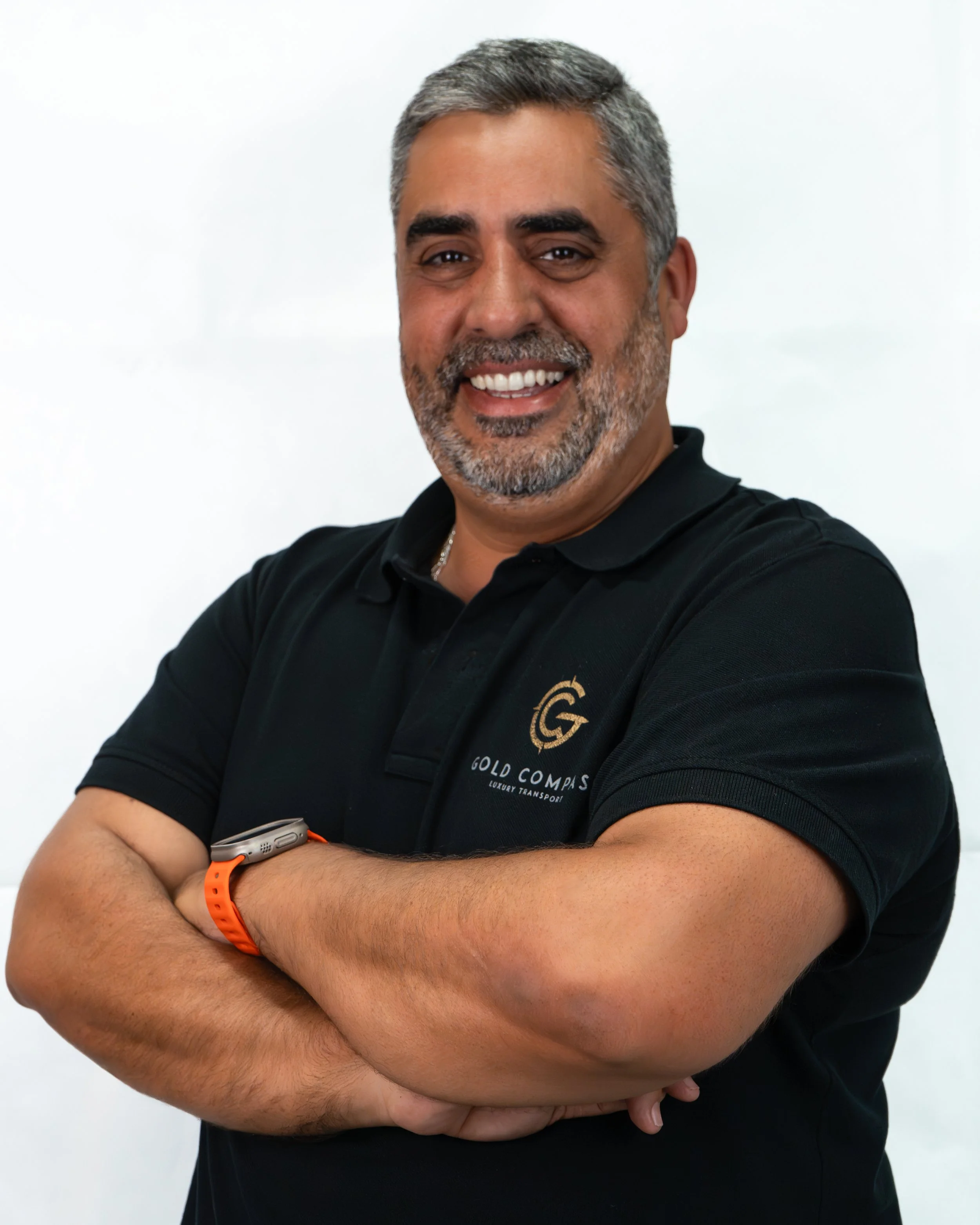 Homem sorridente de cabelo acinzentado, barba, com braços cruzados usando camisa preta com logo e texto 'Gold Compass Luxury Transport', acessórios de relógio e pulseira laranja, posando contra fundo branco.