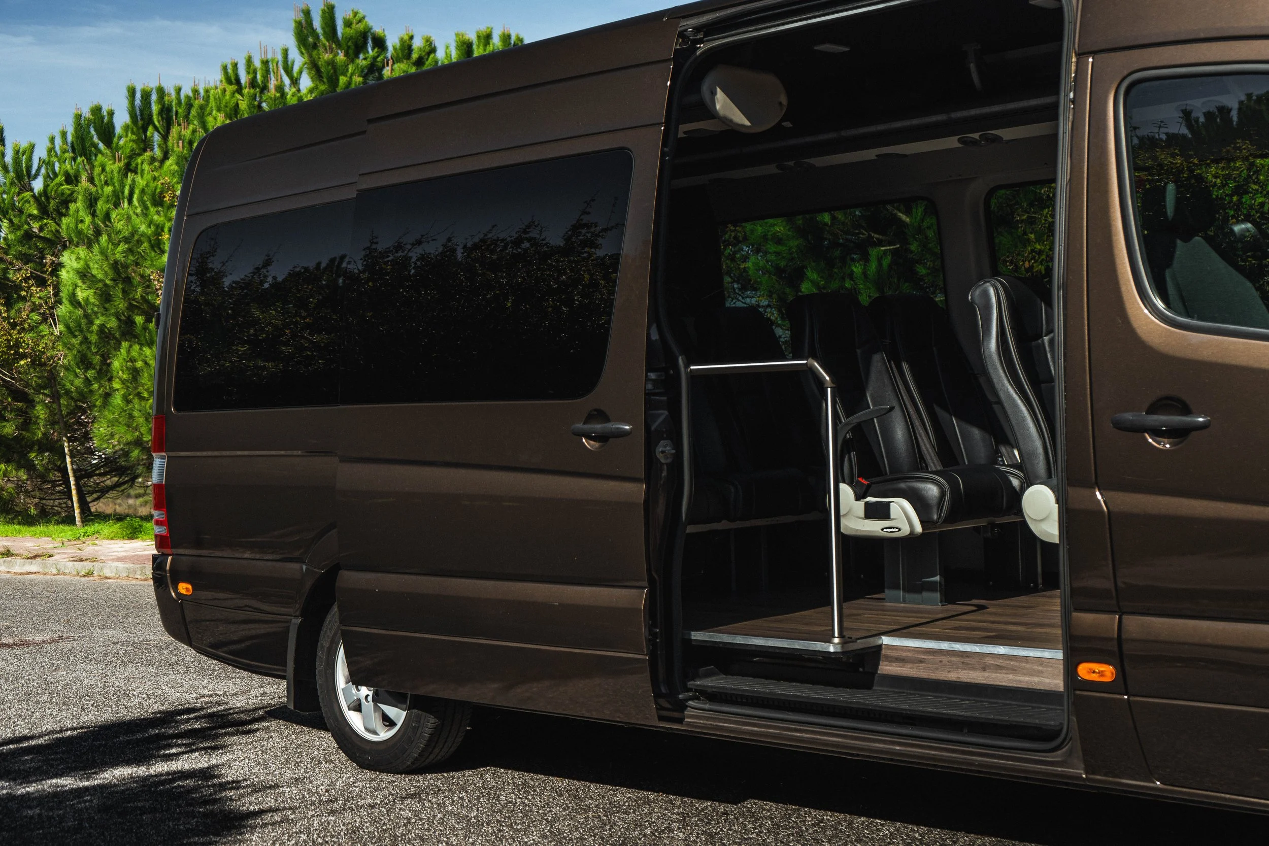 Van preta com portas abertas, interior com assentos de couro e espaço adaptado para cadeira de rodas, estacionada na rua com árvores ao fundo.