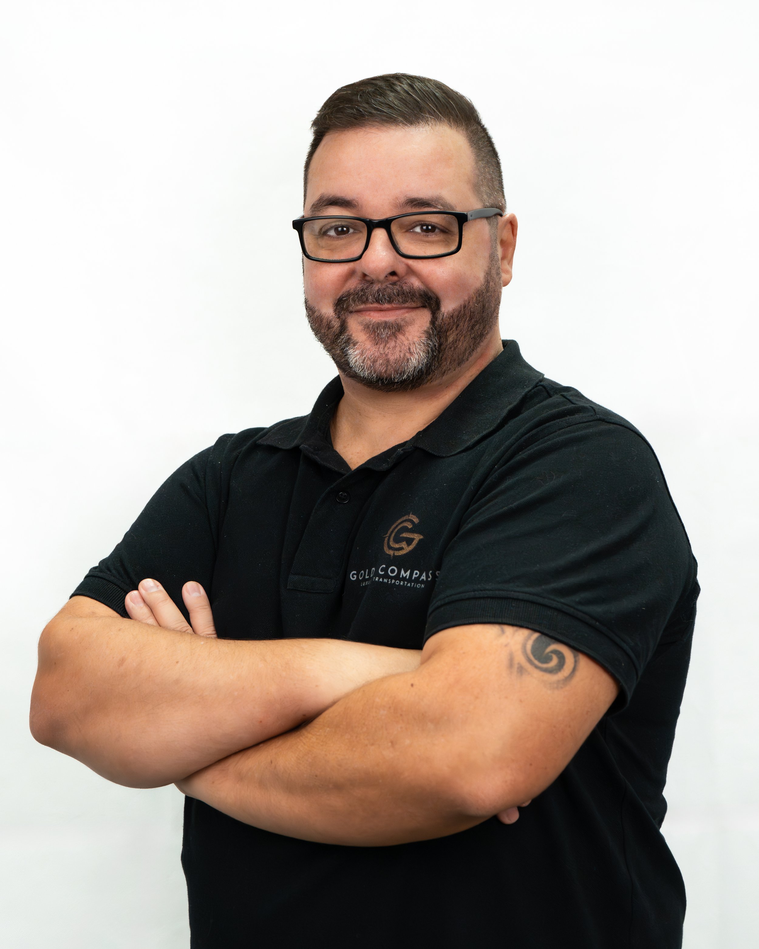Homem sorridente com barba, óculos pretos e camiseta preta com logotipo da Gold Compass, cruzando os braços, contra fundo branco.