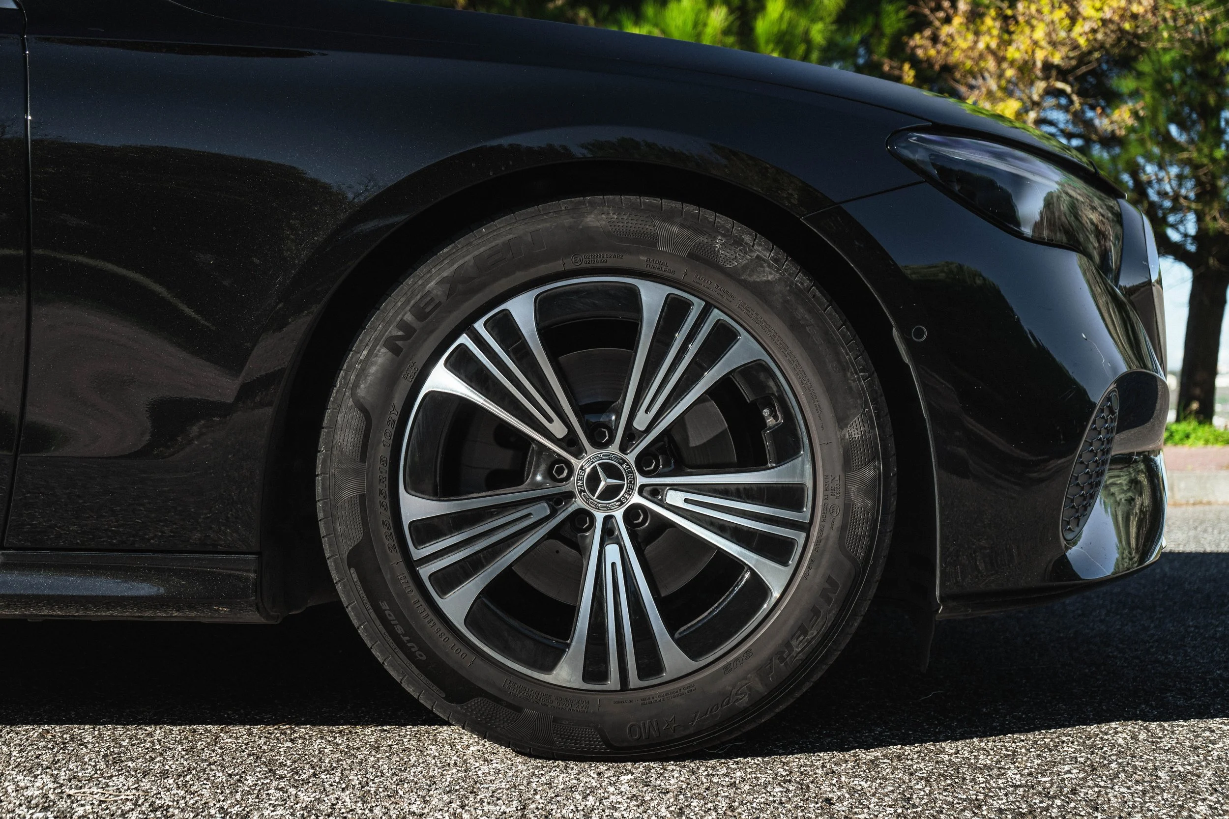 Frontal da roda de um carro preto, com detalhe da marca Mercedes-Benz e pneu Continental, estacionado em uma rua com árvores ao fundo.