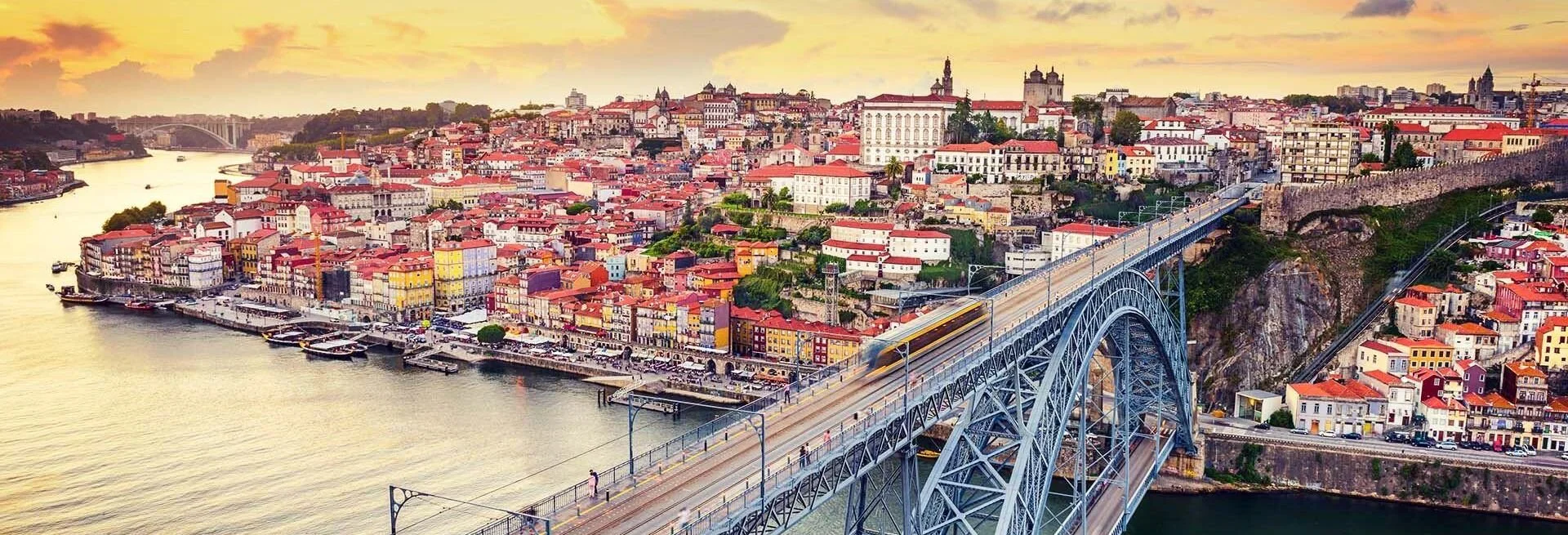 Cidade colorida com rio e ponte em primeiro plano, edifícios históricos ao fundo, ao pôr do sol.