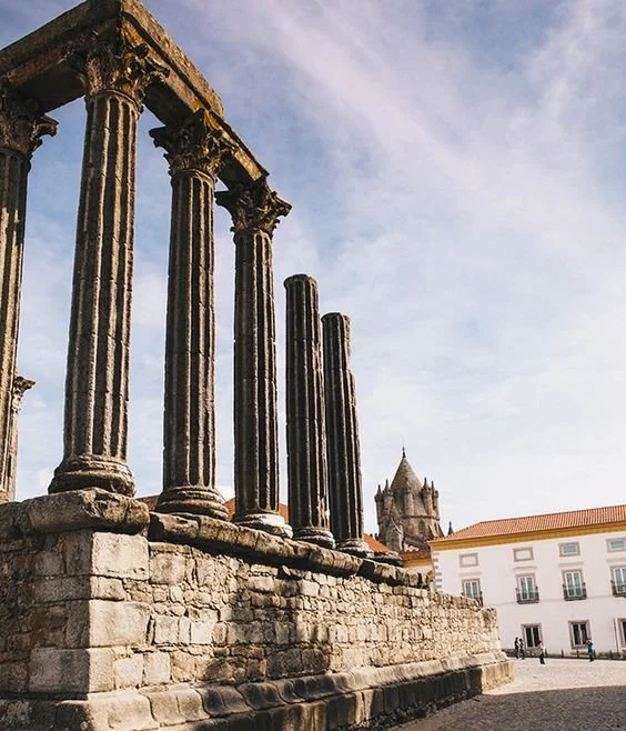 Restauradas colunas antigas de pedra, parte de uma estrutura clássica, com um prédio e uma torre ao fundo, sob céu parcialmente nublado.
