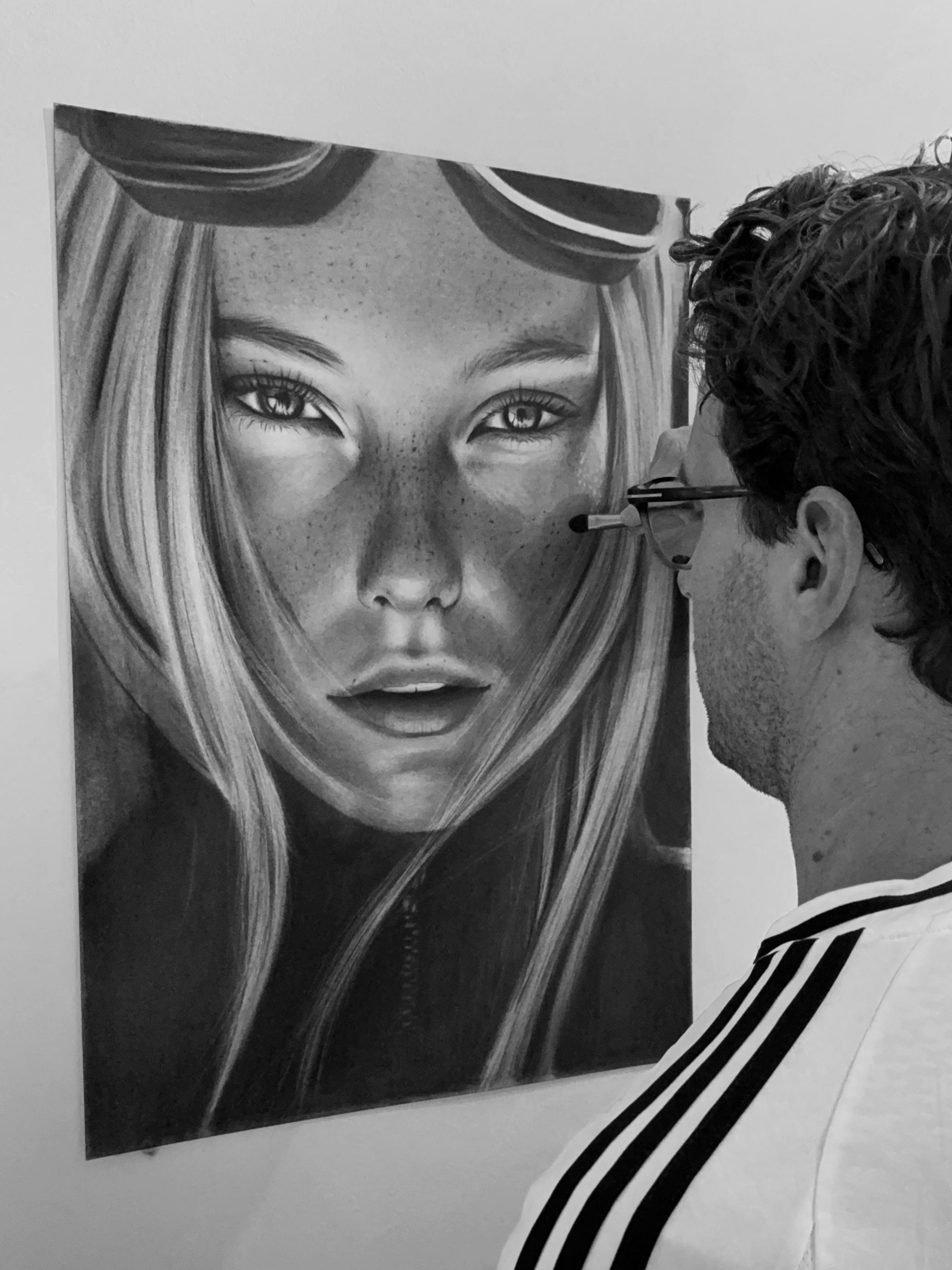 Photograph of an artist painting a realistic black and white portrait of a woman on a canvas, showing the artist's face in profile and the detailed artwork of the woman's face with long hair and freckles.