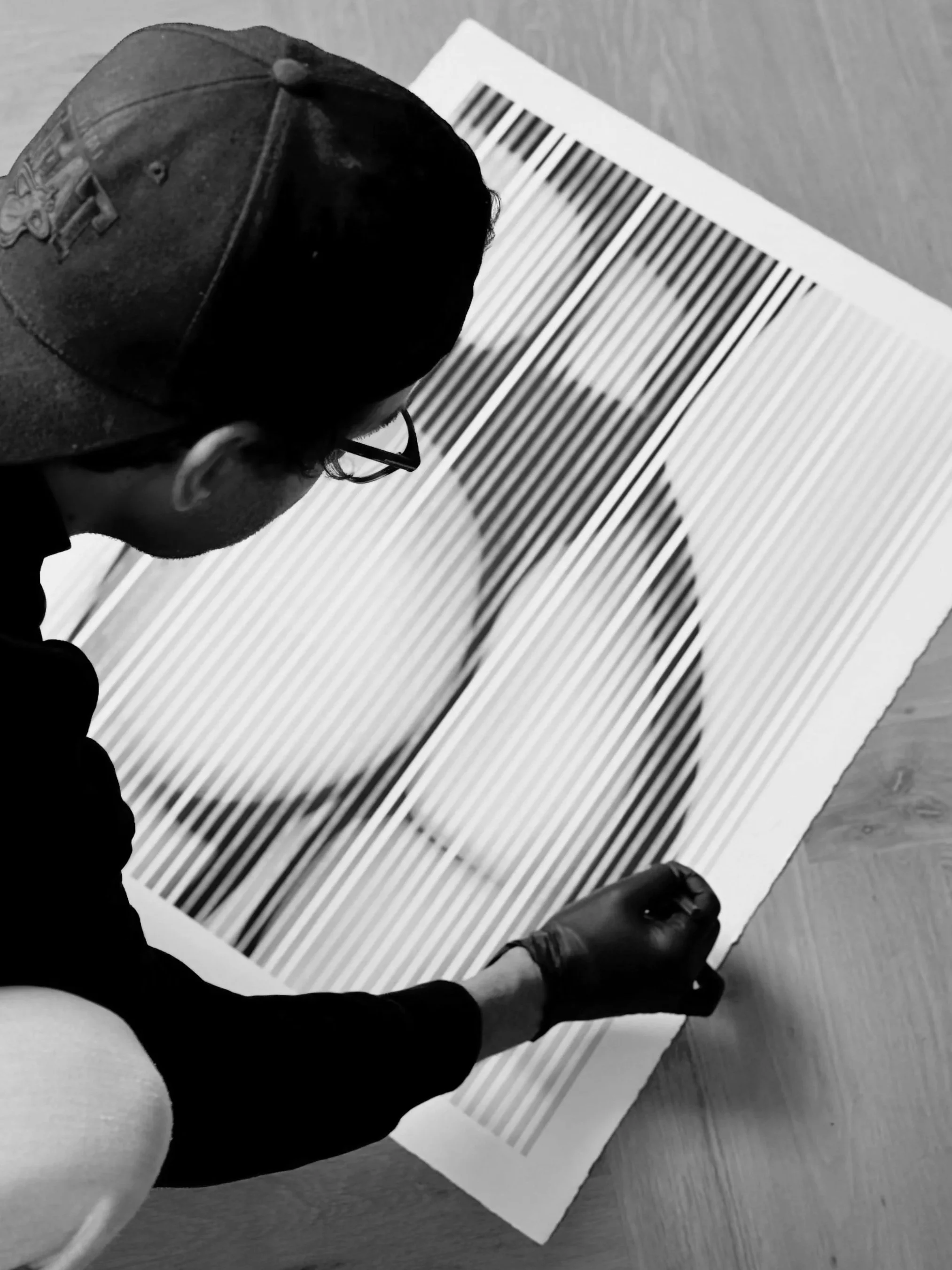 Person wearing a cap and glasses using a glue stick on a striped sheet of paper, viewed from above.
