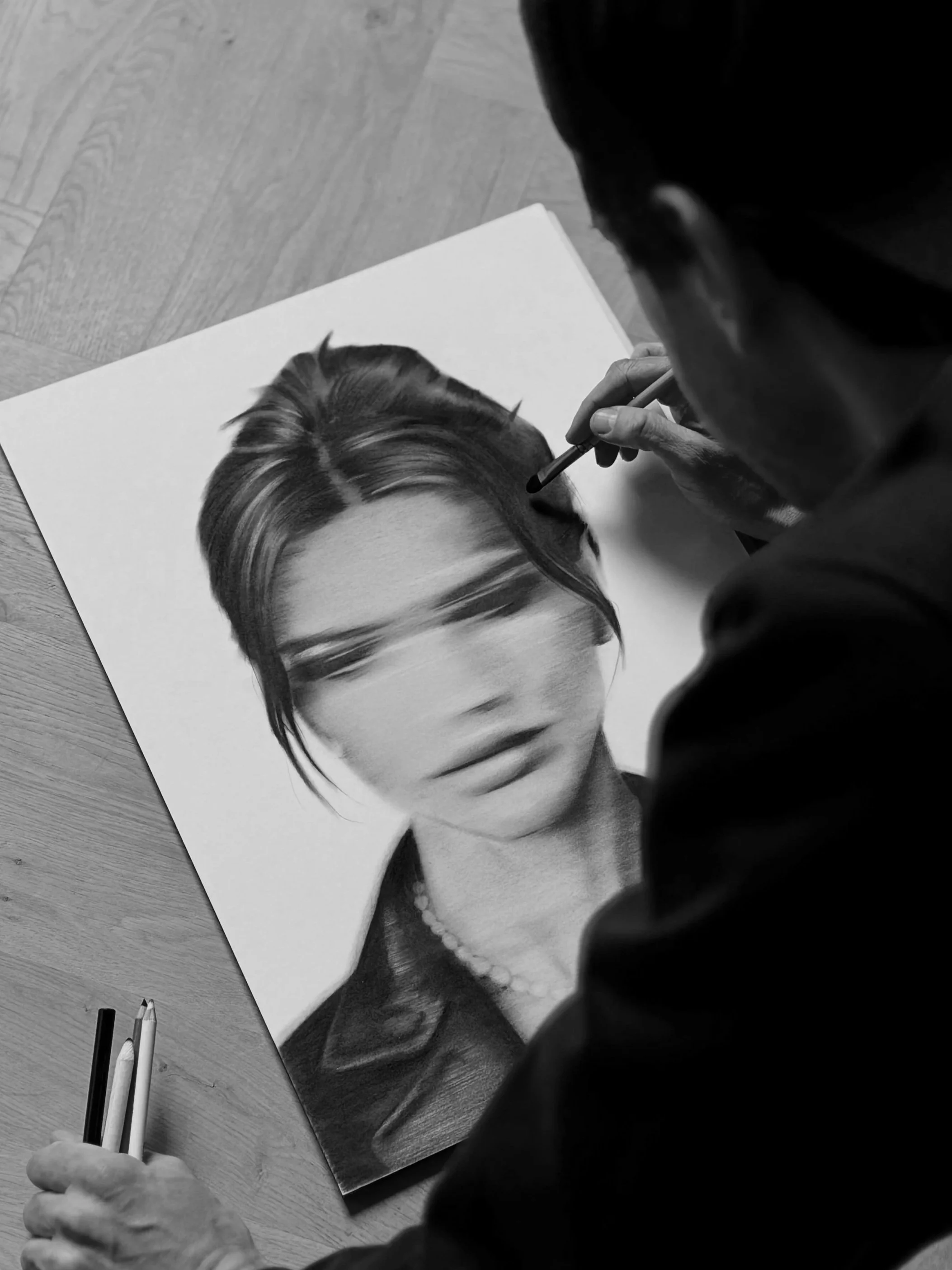 A person drawing a realistic black and white portrait of a woman with short hair and a serious expression, using colored pencils on a large sheet of paper placed on a wooden surface.