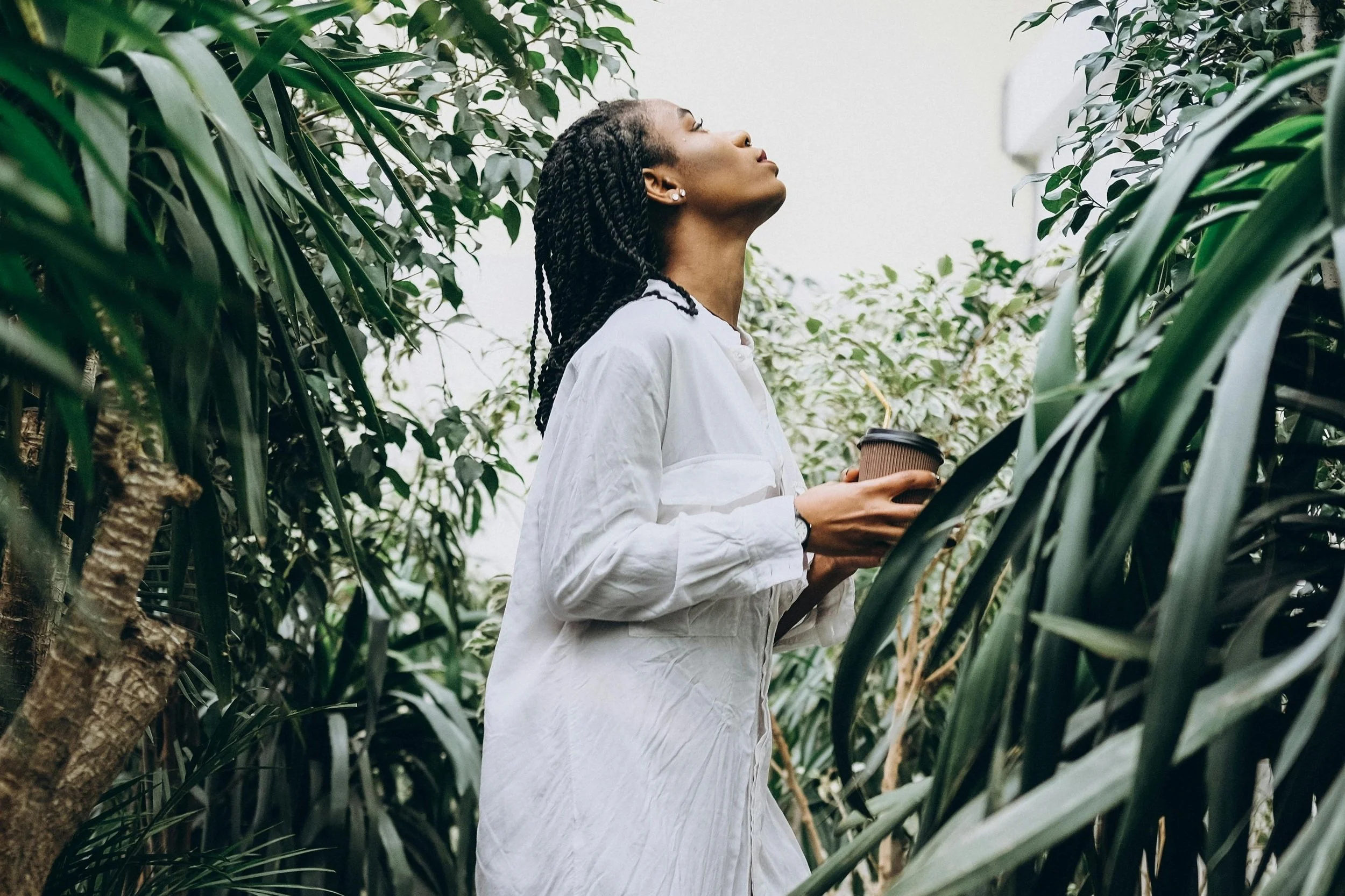 A woman in a white shirt holding a coffee cup, standing among lush green plants, with her head tilted back and eyes closed.