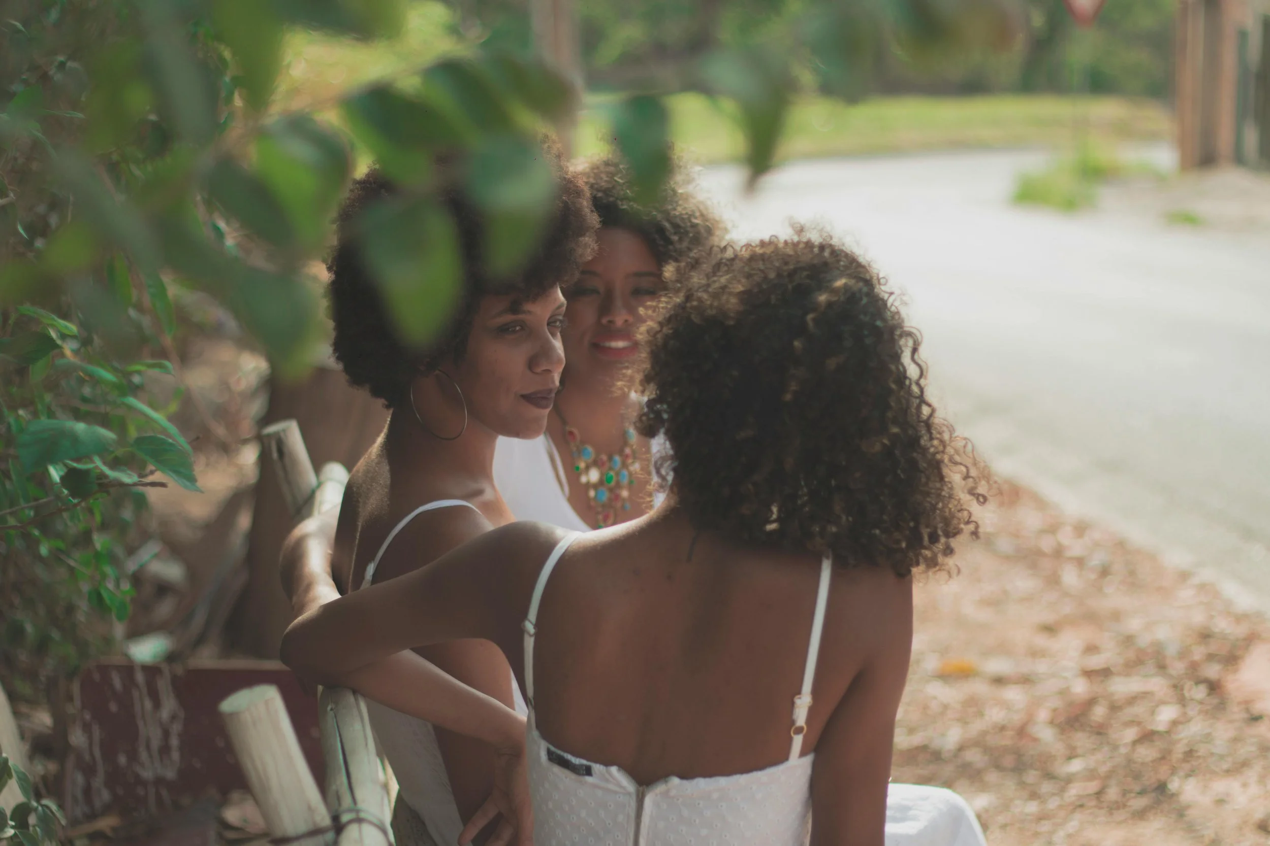 Three women with curly hair sitting on a park bench, engaged in conversation, framed by green leaves, with a quiet street in the background.