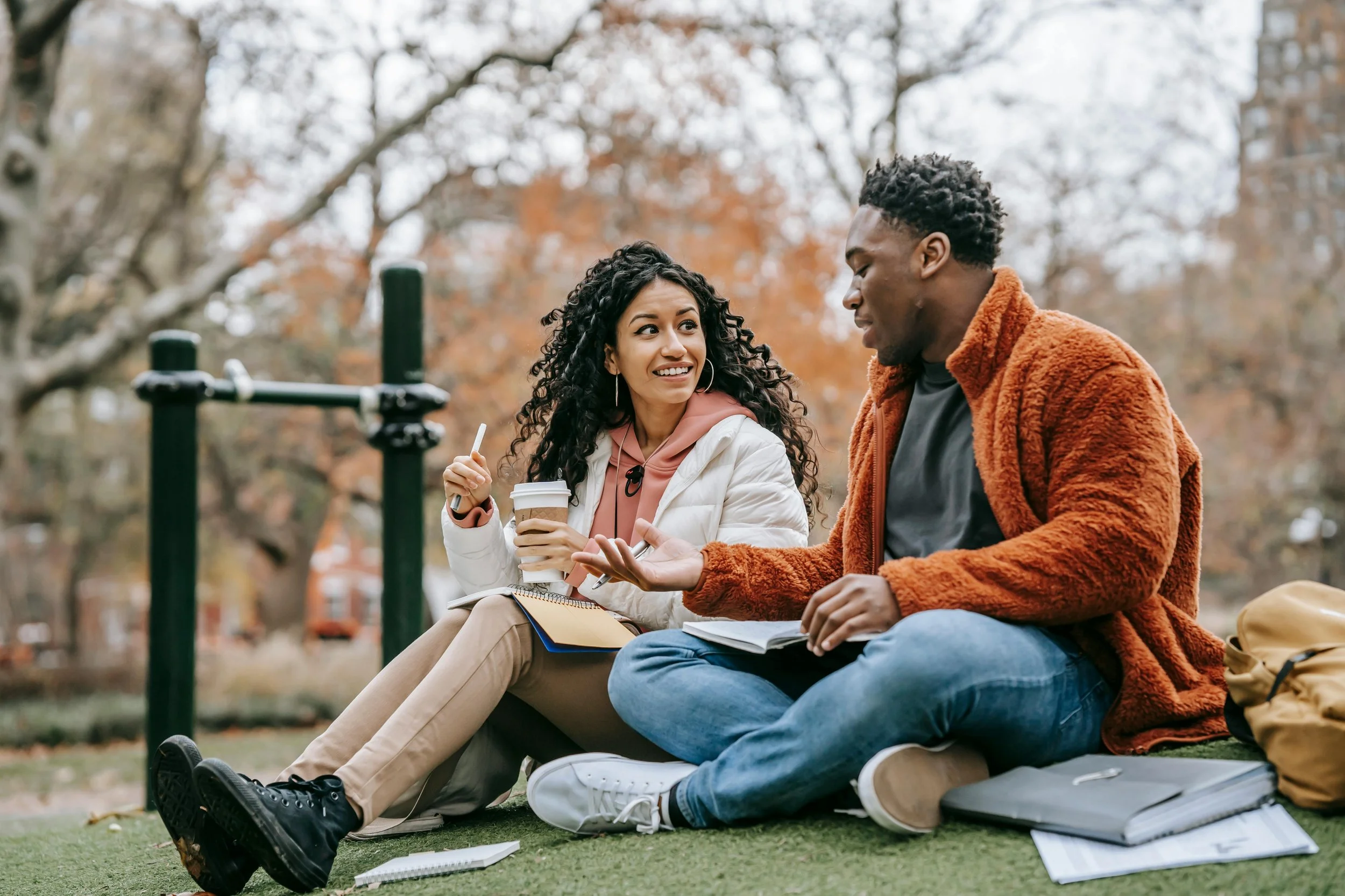 A young woman and man sitting on grass in a park, engaging in a friendly conversation, surrounded by autumn trees and buildings in the background. They have notebooks, a laptop, and coffee cups with straws.