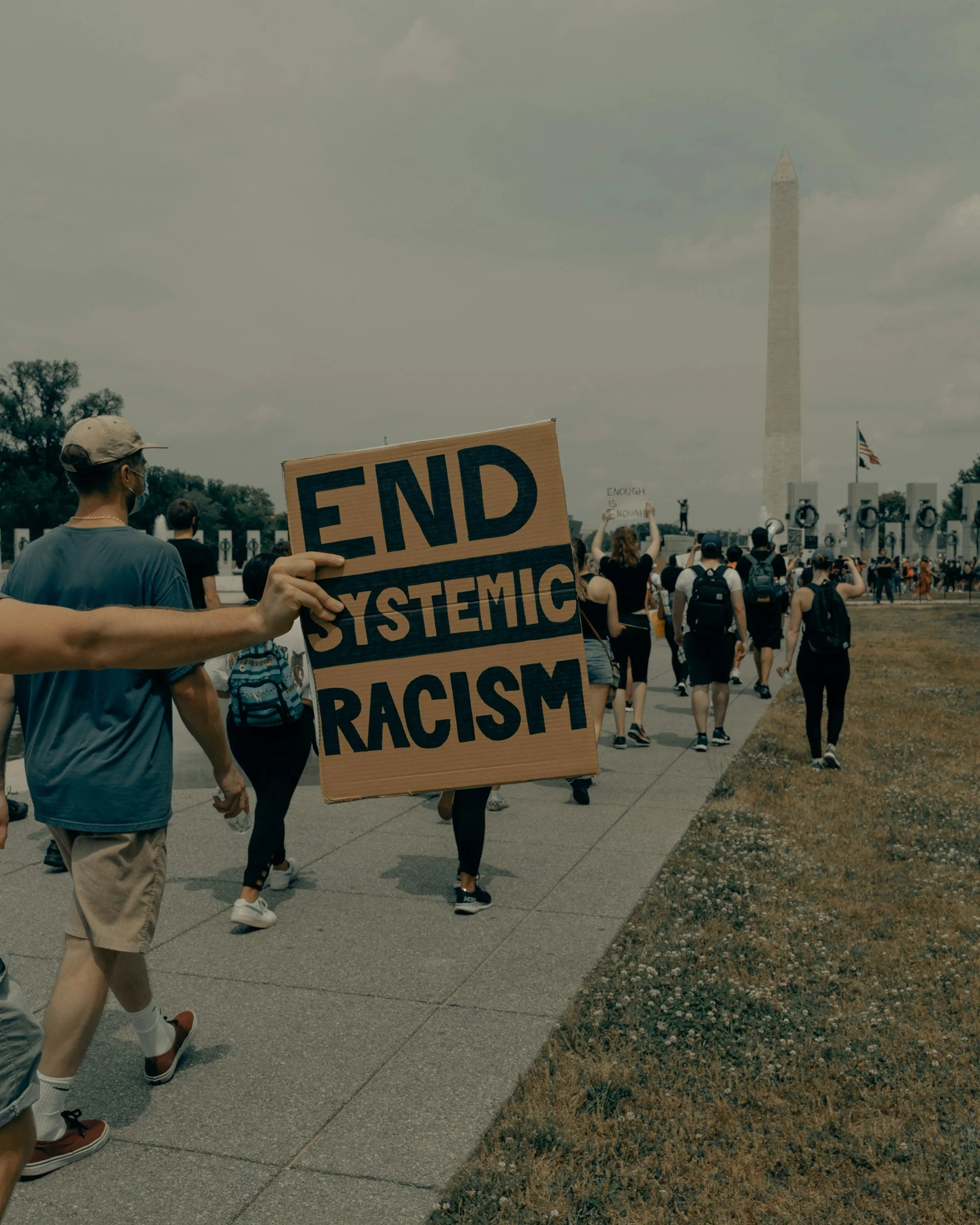 People participating in a protest march near the Washington Monument, holding signs including one that reads "End Systemic Racism." The scene is outdoors on a cloudy day, with a crowd walking towards the monument and some individuals taking pictures.