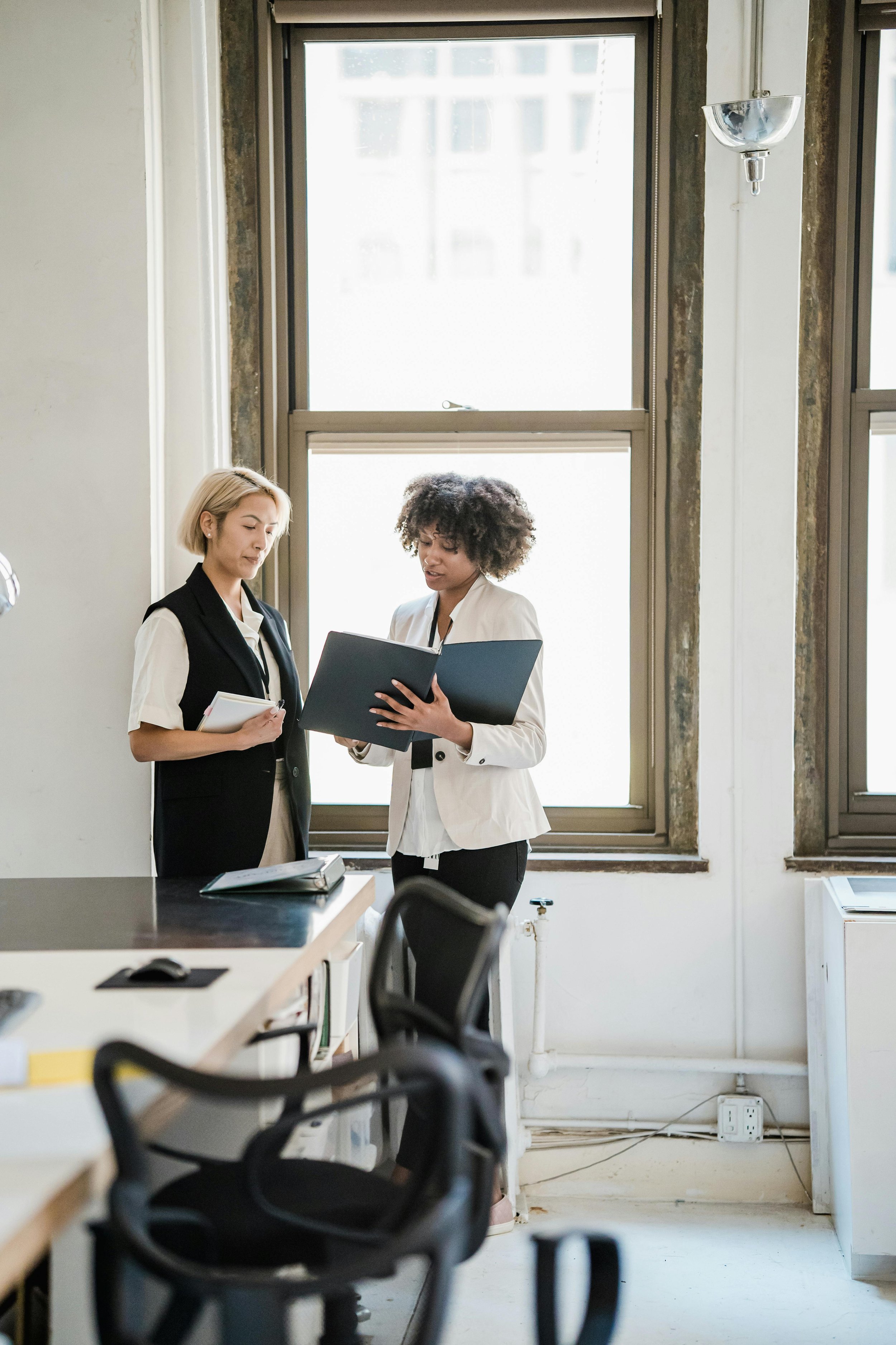 Two women standing and talking in an office with large windows, one holding a notebook and the other a folder, near a table with a notebook and a mouse.