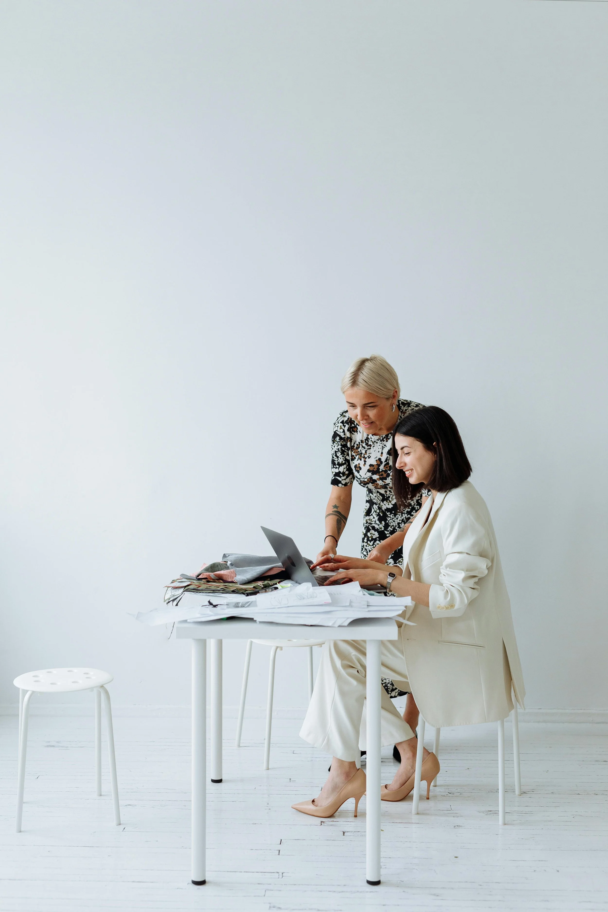 Two women working together at a white table, surrounded by scattered papers and fabric swatches, in a minimalist white room. One is seated in a beige suit with heels, the other stands in a patterned dress.