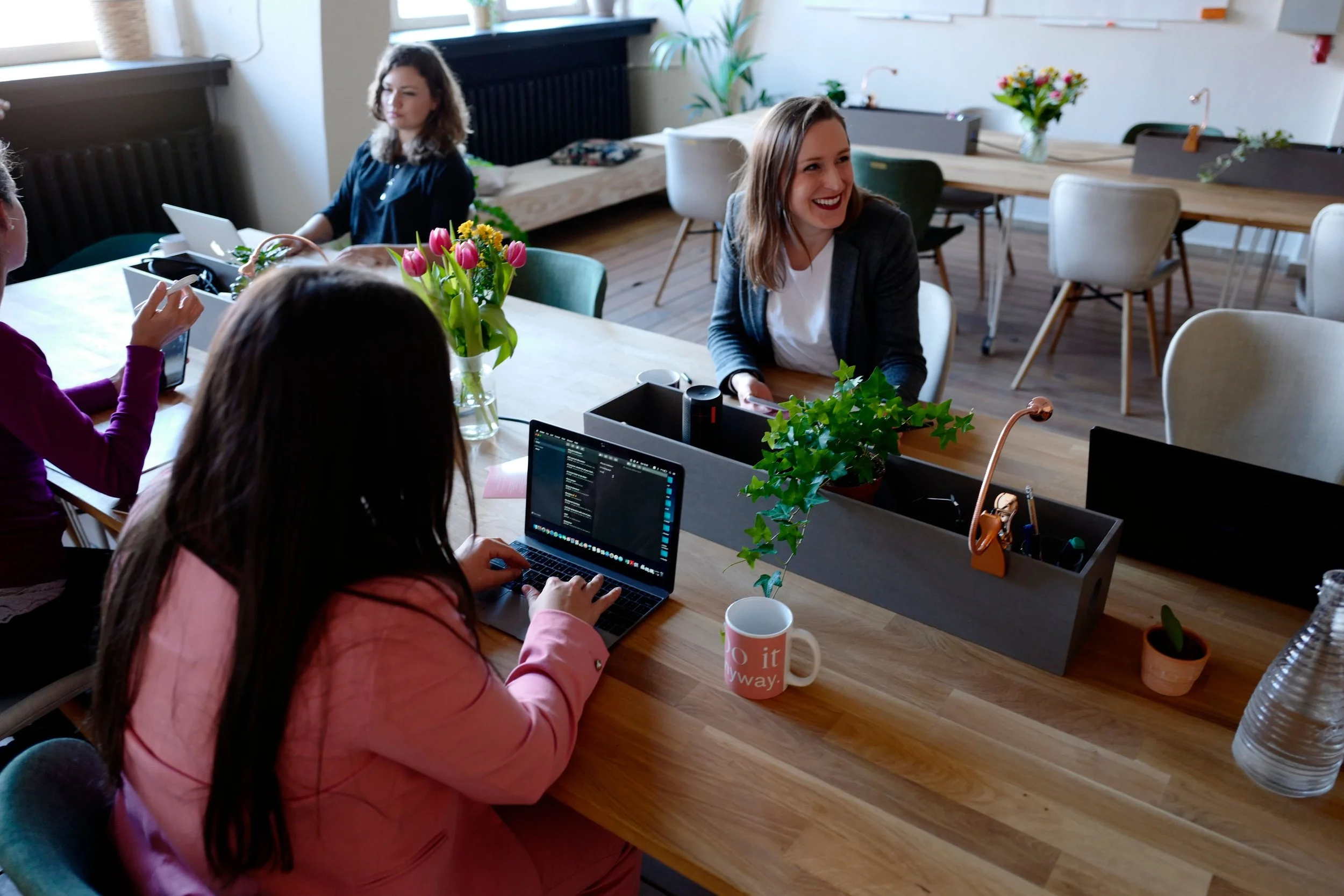 Group of women working together in a bright, modern office space with plants, flowers, laptops, and coffee mugs on a wooden table.