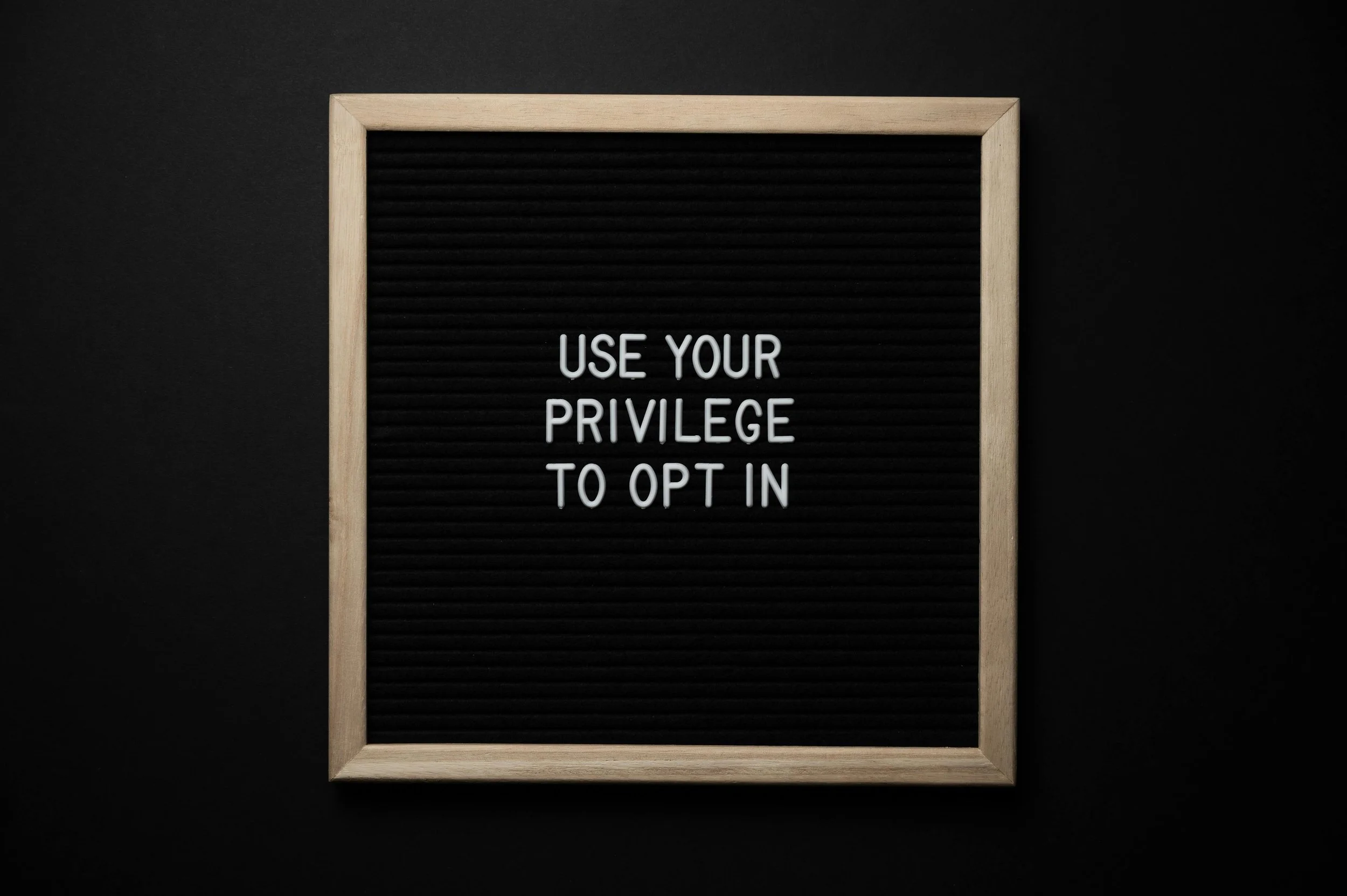 Black letter board with light wood frame displaying the message 'Use your privilege to opt in' in white letters.