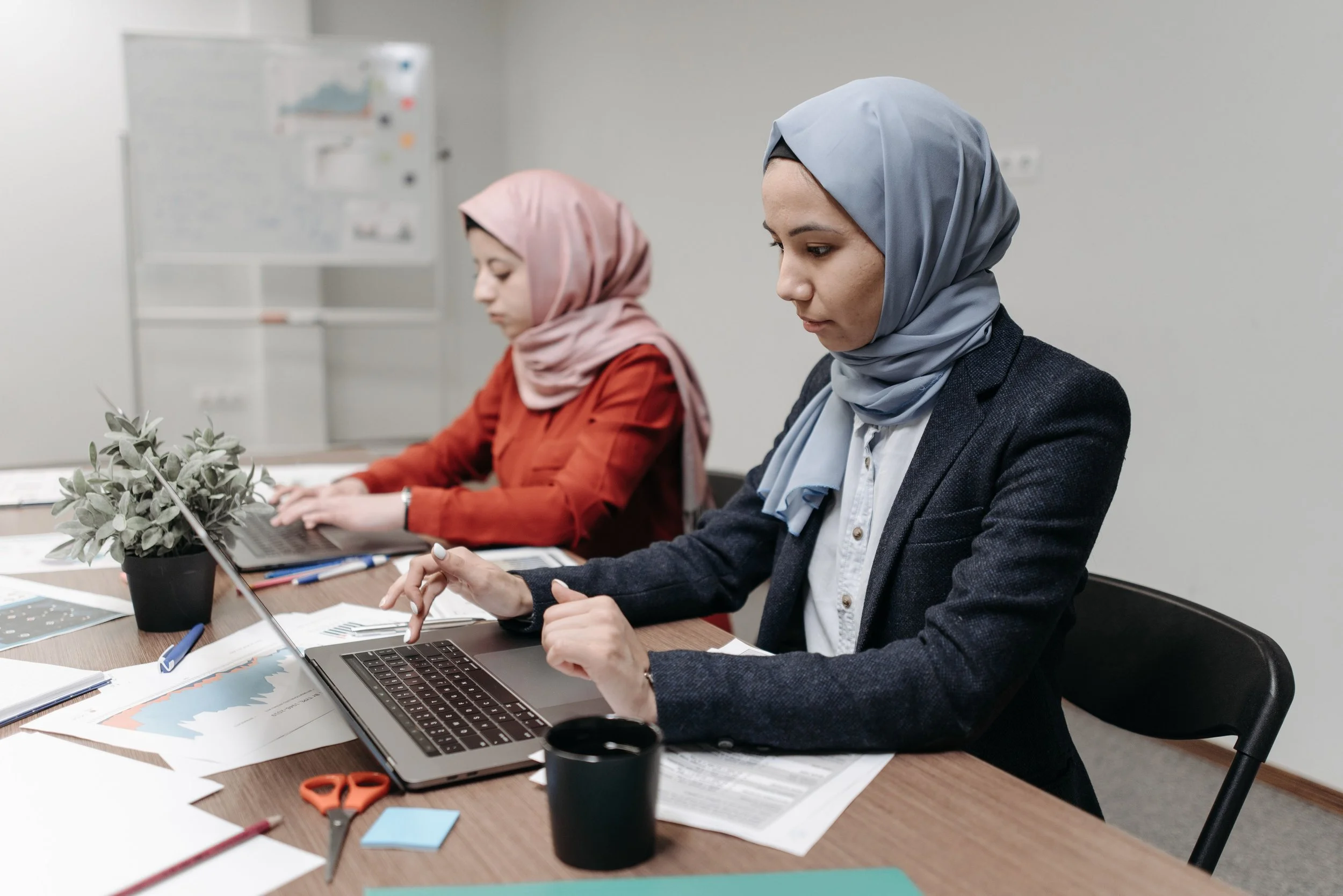 Two women wearing headscarves working on laptops at a table in an office setting, with papers, office supplies, and a plant in the foreground.
