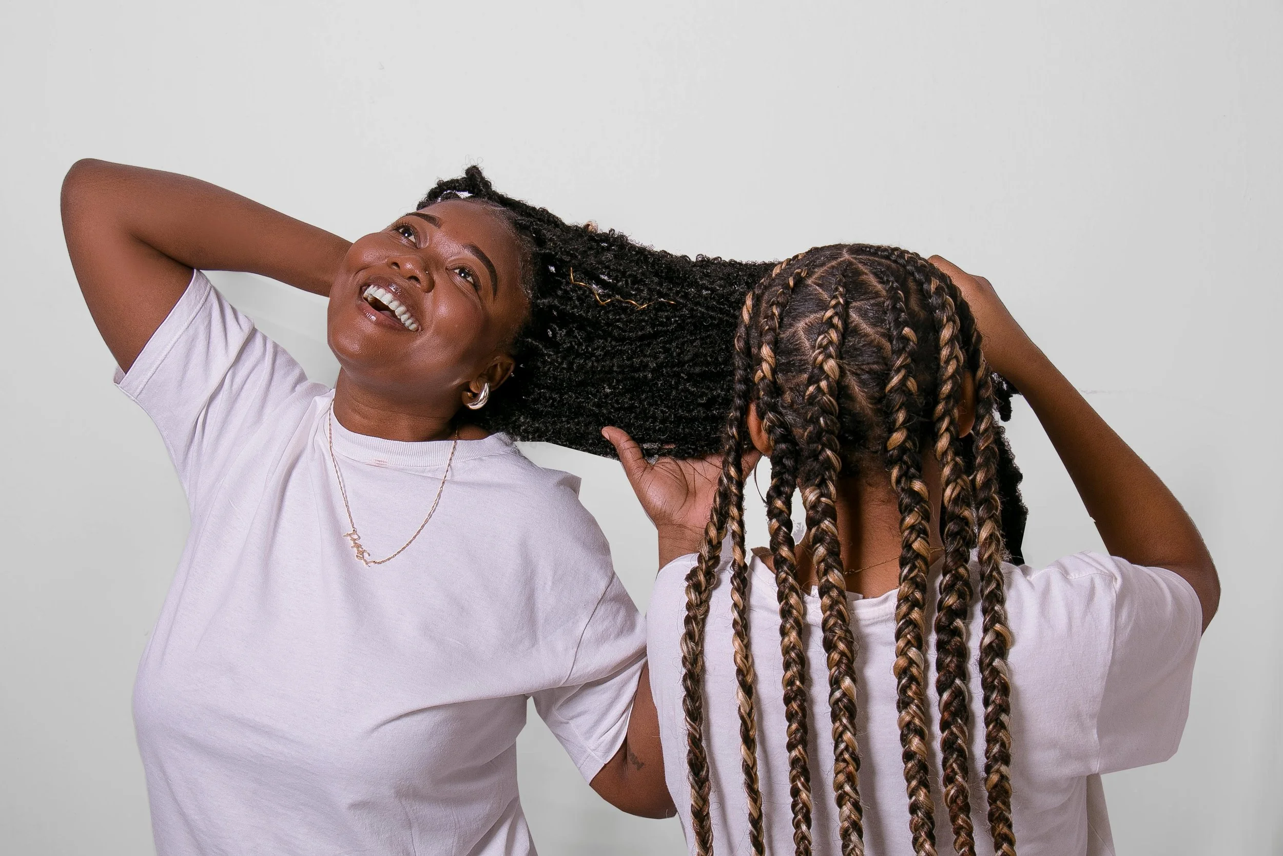A woman with dark skin and long braided hair smiling while adjusting her hair with another woman, also with dark skin, who is seen from behind with braided hair, both wearing white shirts, against a plain white background.