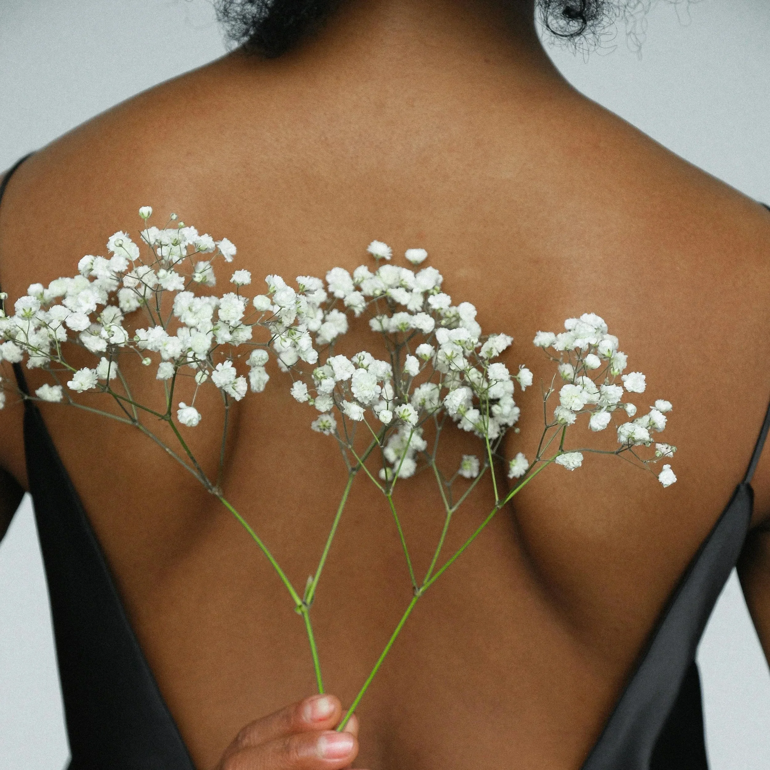 A person with medium brown skin holding a bouquet of white small flowers against their back, with a black dress with spaghetti straps.