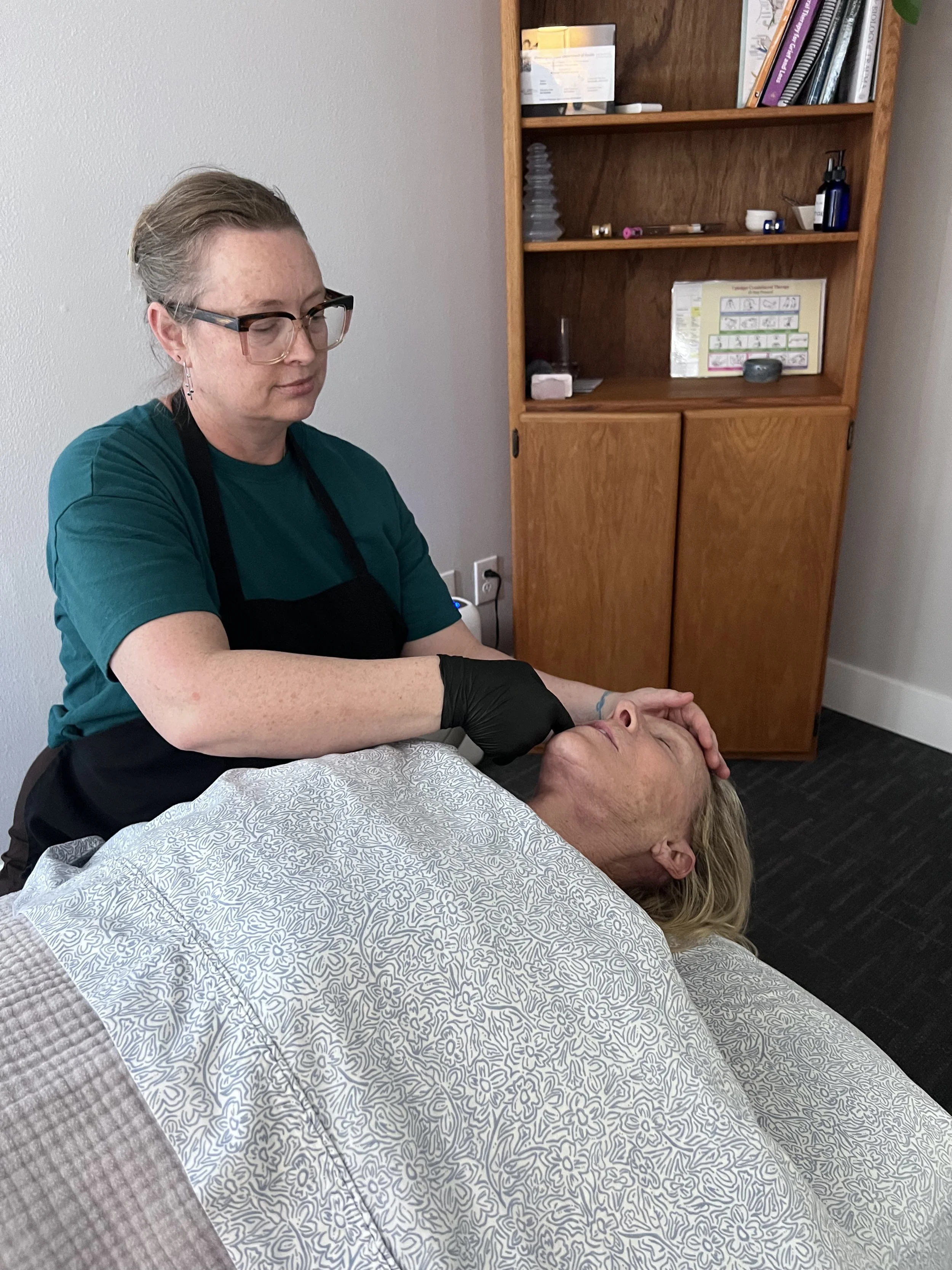 A woman with blonde hair lying on a bed and receiving a facial massage from a therapist with glasses and black gloves in a room with a wooden bookshelf.