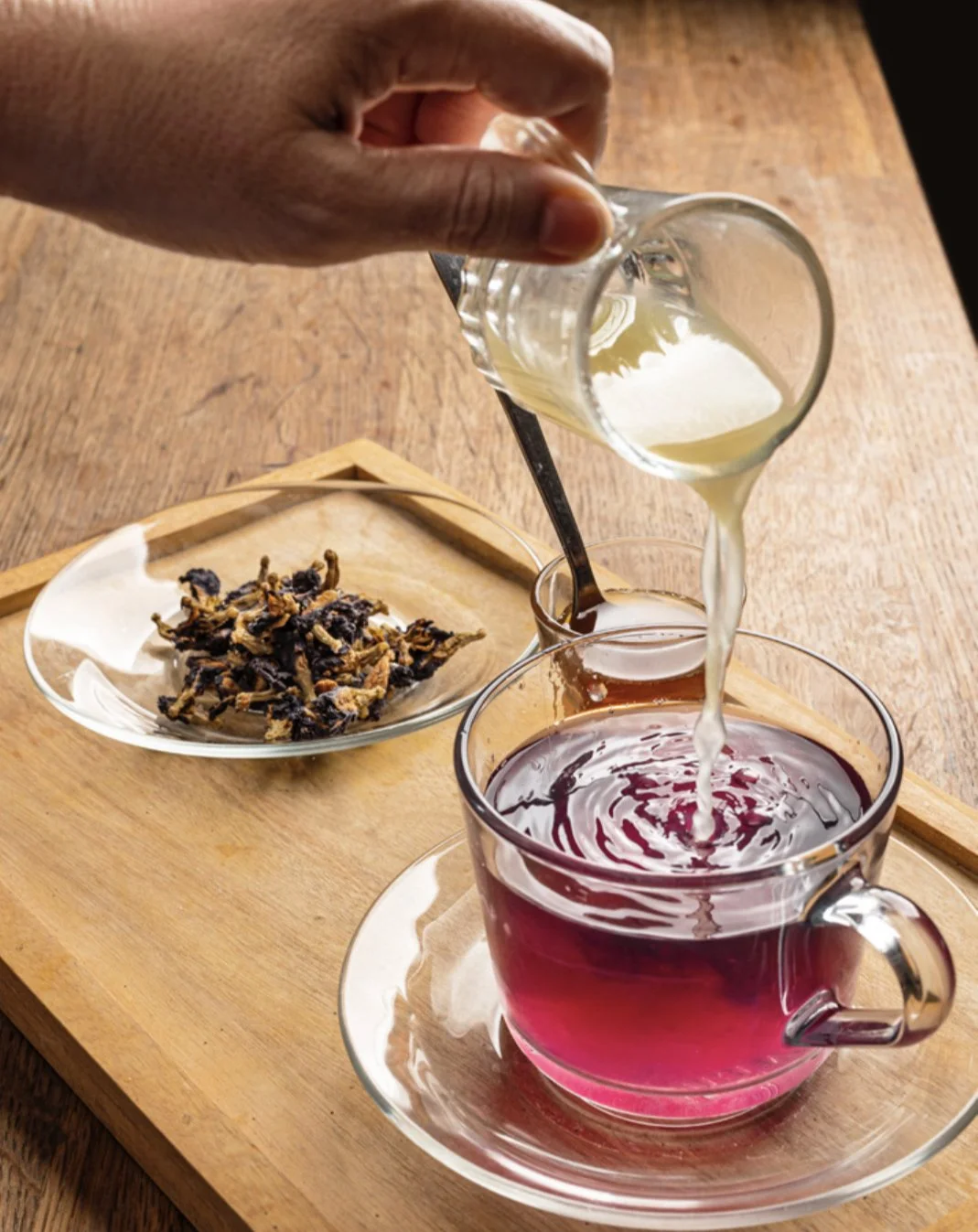 A hand pouring cream into a cup of red herbal tea that is on a wooden tray. In the background, a plate with dried herbs or flowers.