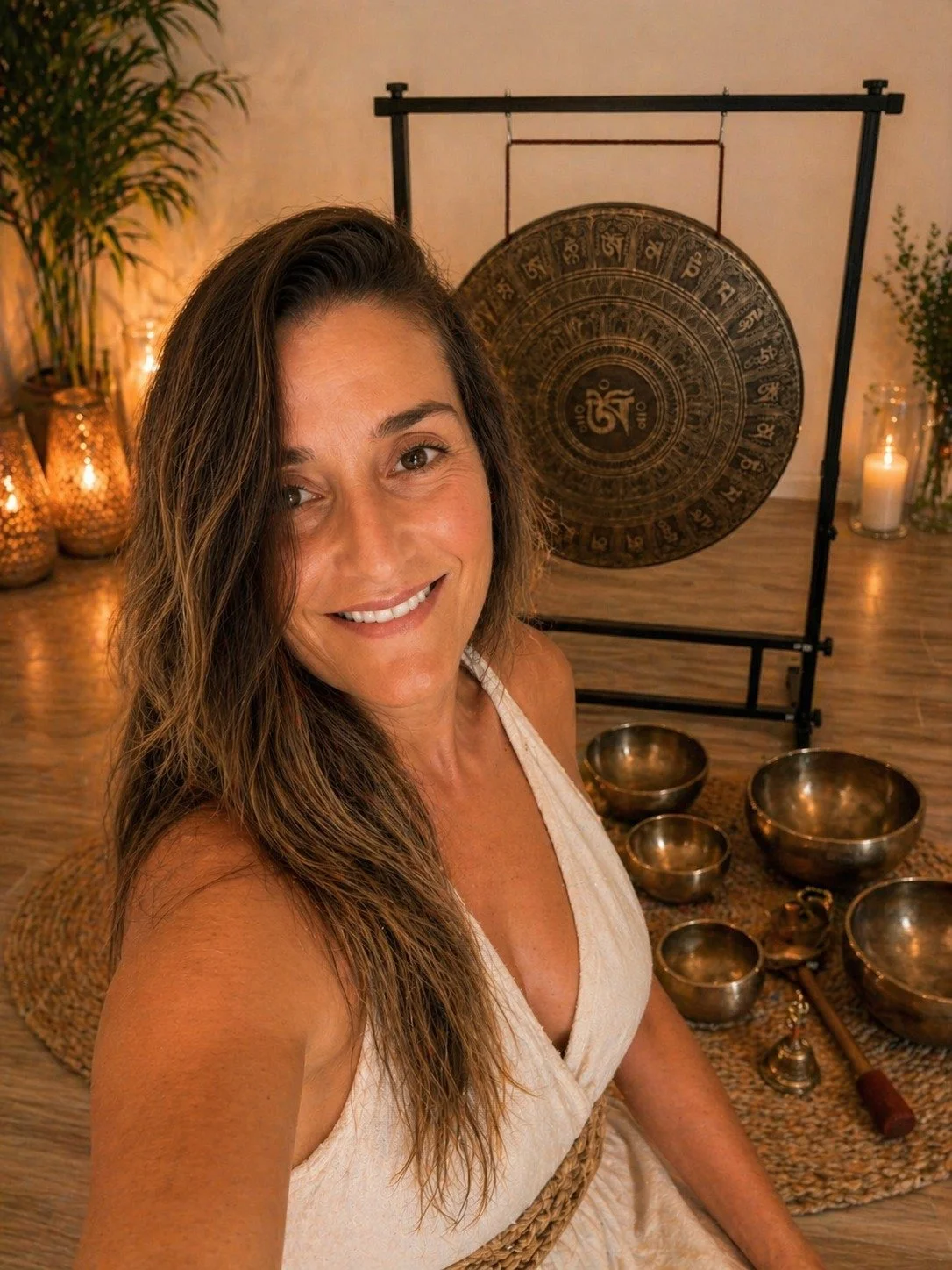 A woman smiling in front of a gong and Tibetan singing bowls setup, with candles and plants in the background.