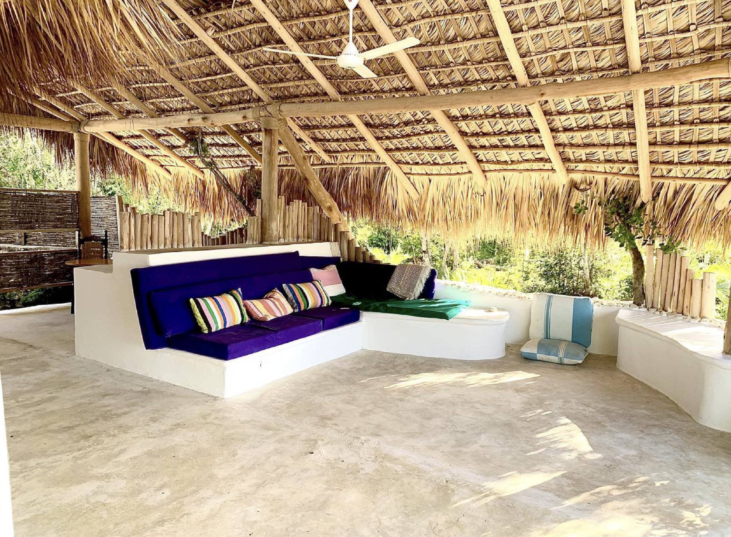 Open-air lounge area with colorful cushions and a thatched roof, surrounded by greenery.