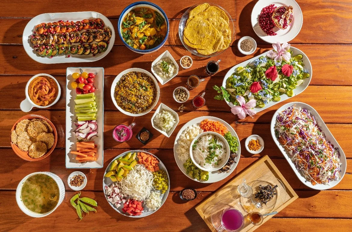 A variety of traditional Mexican dishes and drinks are spread out on a wooden table, including tacos, salads, sauces, and desserts.