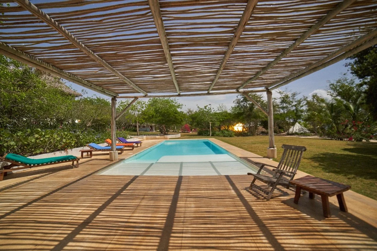 Swimming pool with lounge chairs under a bamboo shade, surrounded by greenery and trees at sunset.