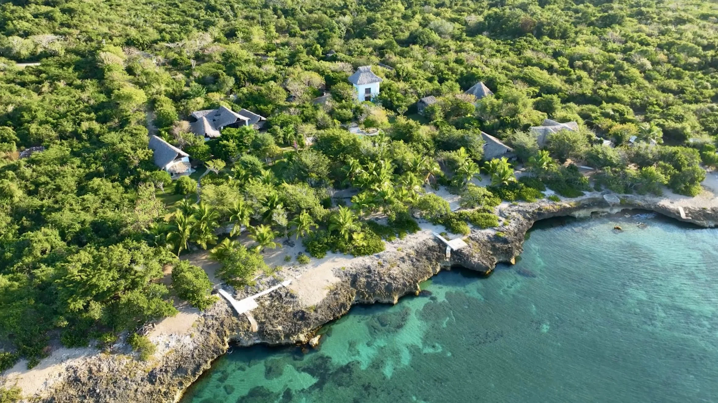 Aerial view of a tropical beach with rocky shoreline, clear blue water, dense green vegetation, and thatched-roof buildings in the jungle.