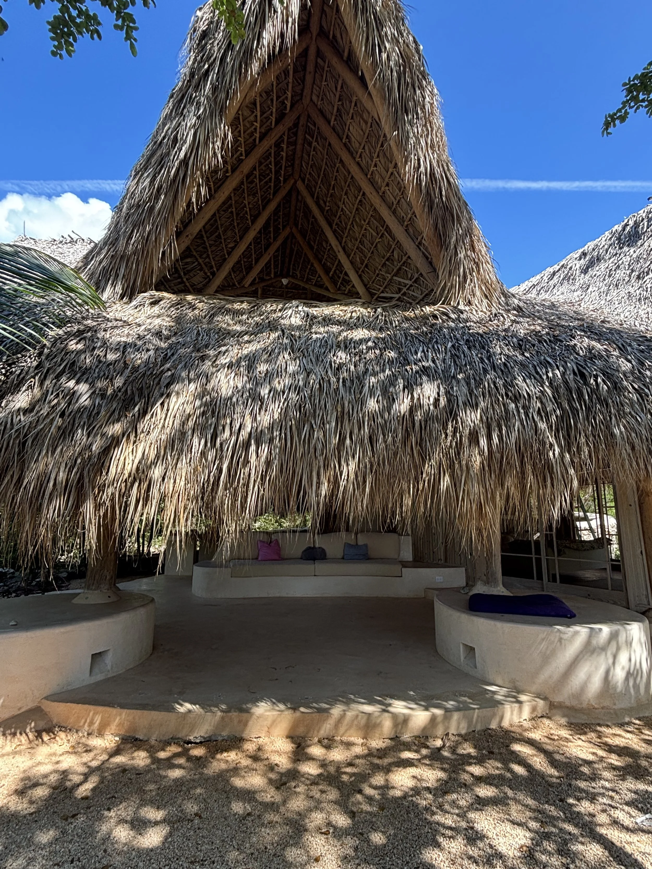 Open-sided hut with a thatched roof, built with wooden beams. Inside, there is a curved white bench with colorful pillows under the shaded area. Shadows from leaves create patterns on the ground, and the sky is clear with a few clouds.