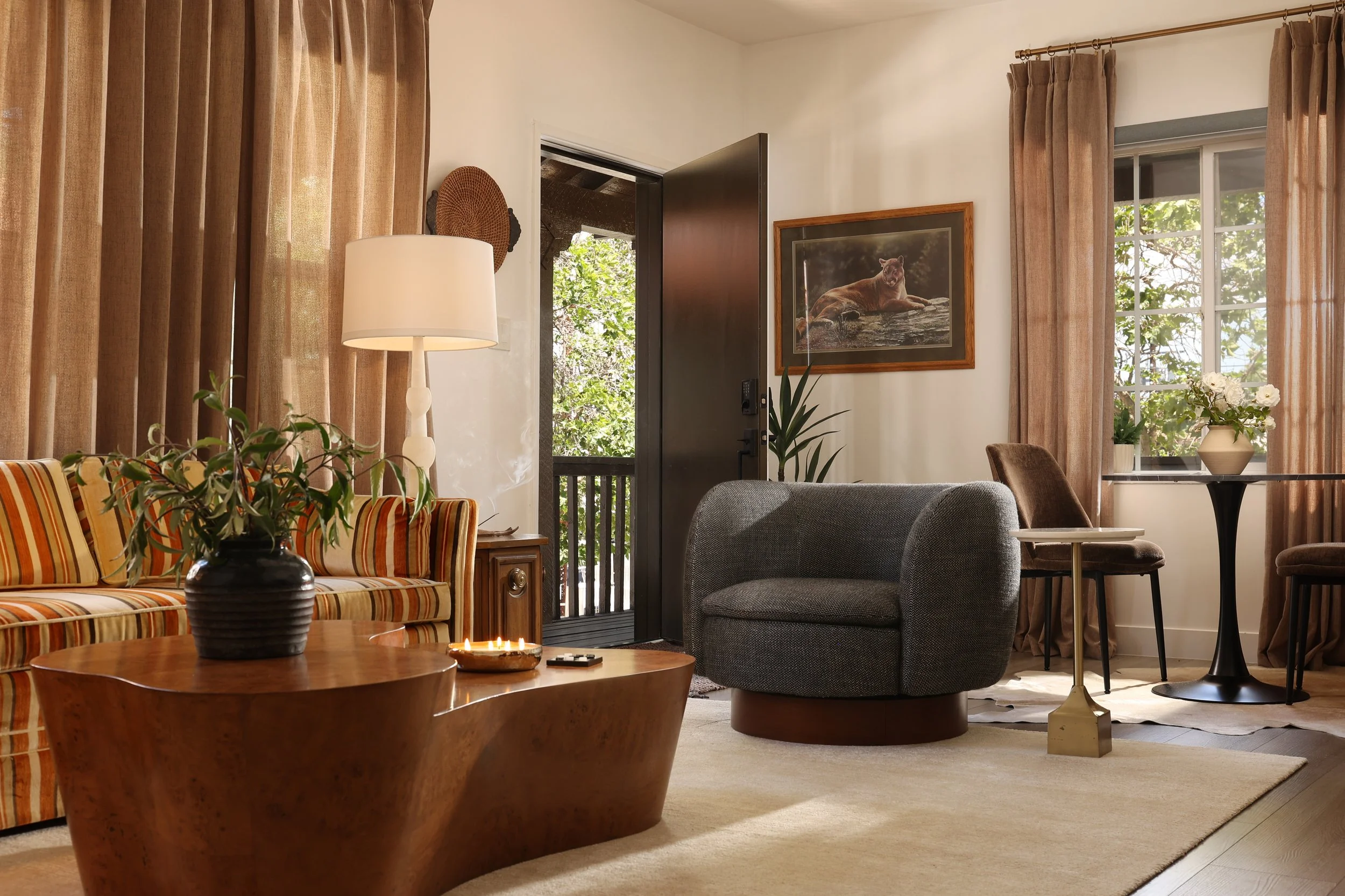 Moody organic modern apartment living room and dining area in Orange, California featuring a vintage striped sofa, marble pedestal dining table, graphite swivel chair, burl wood coffee table, warm wood tones, and Spanish-style architecture.