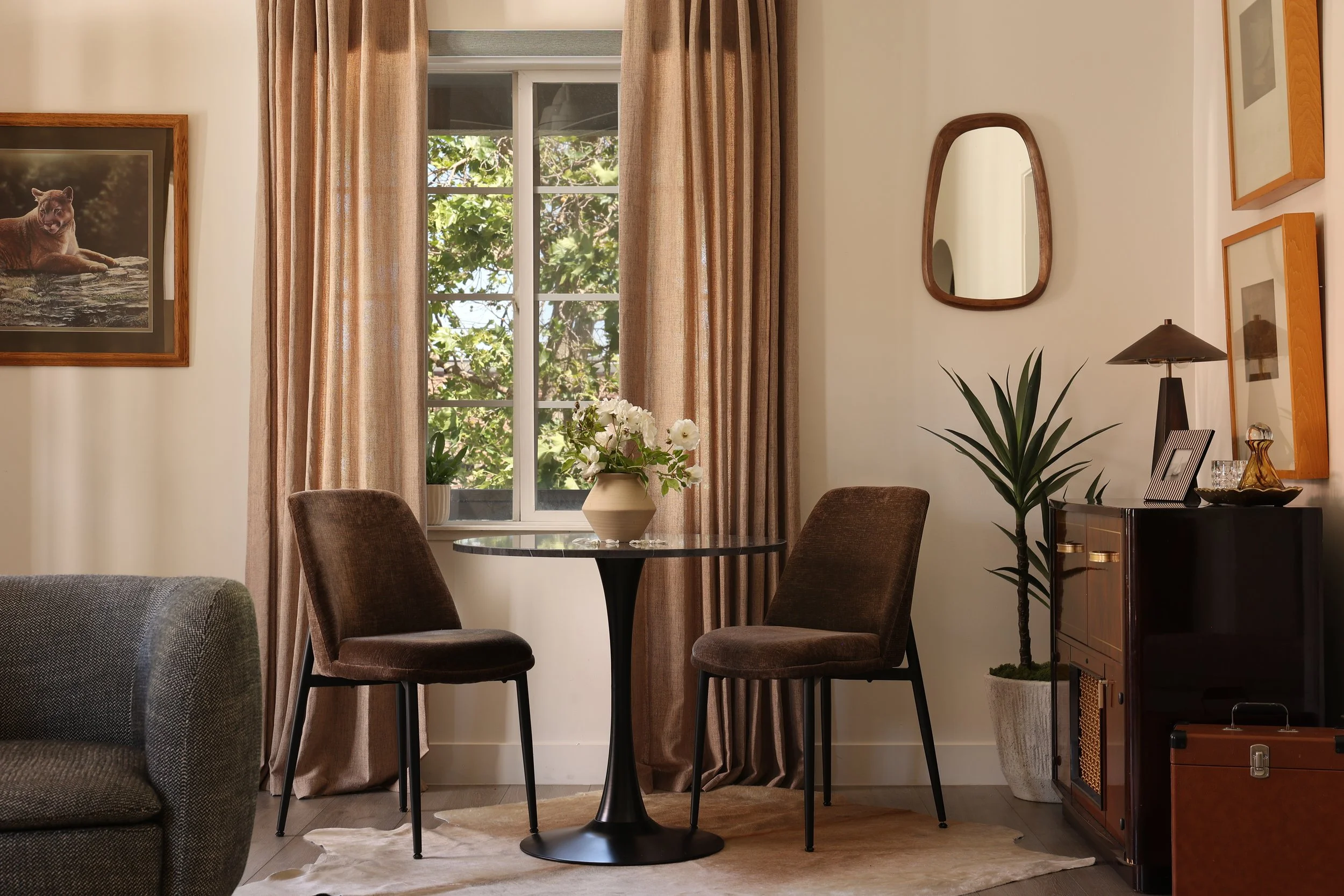 Dining area in a warm moody apartment featuring a cowhide rug, velvet dining chairs, vintage-style record player console, original art, organic mirror, layered greenery, and collected Spanish modern interiors in Orange, California.