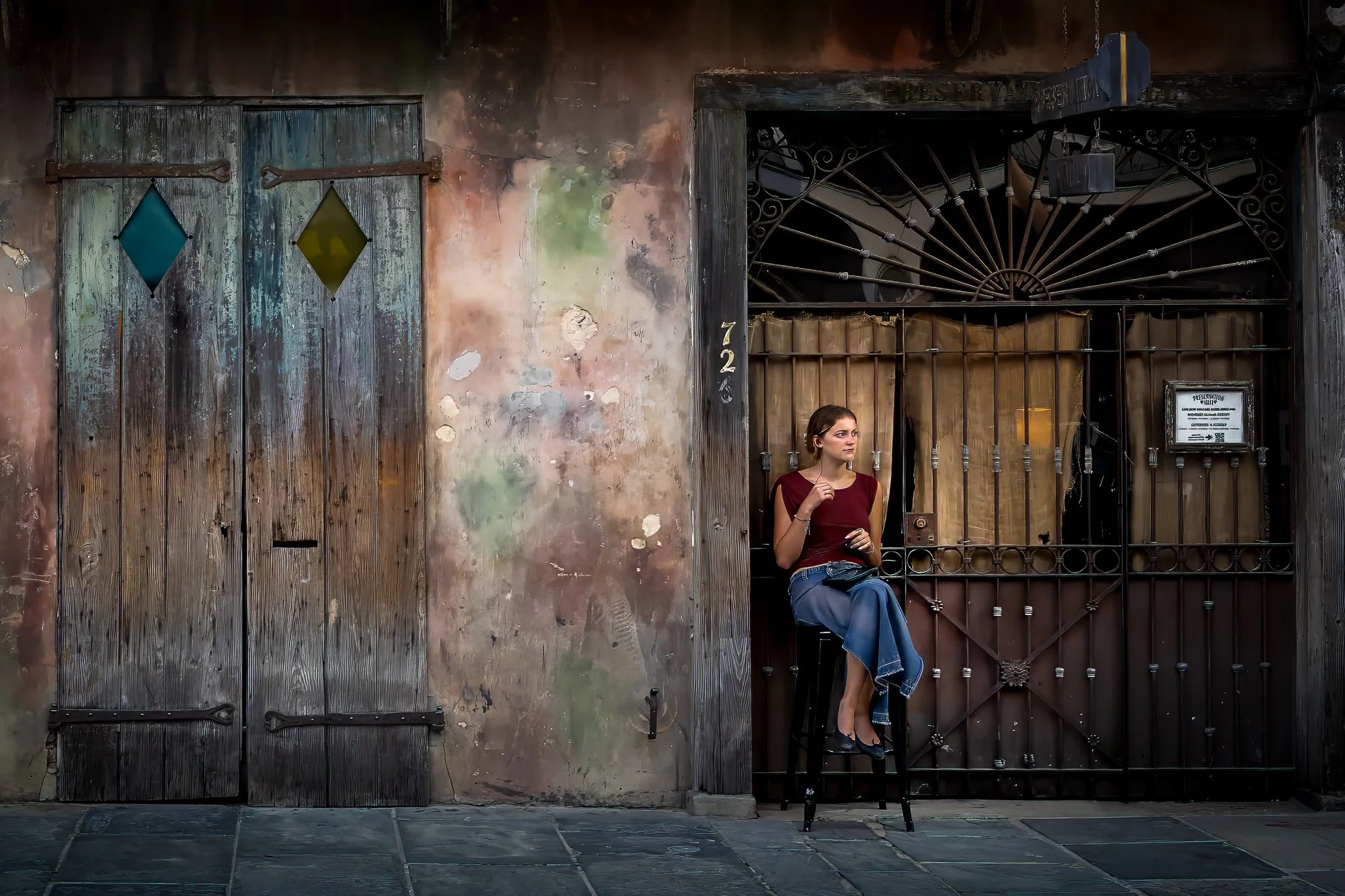 A woman sits outside Preservation Hall in New Orleans beside weathered shutters, old plaster, and ironwork along St. Peter Street.