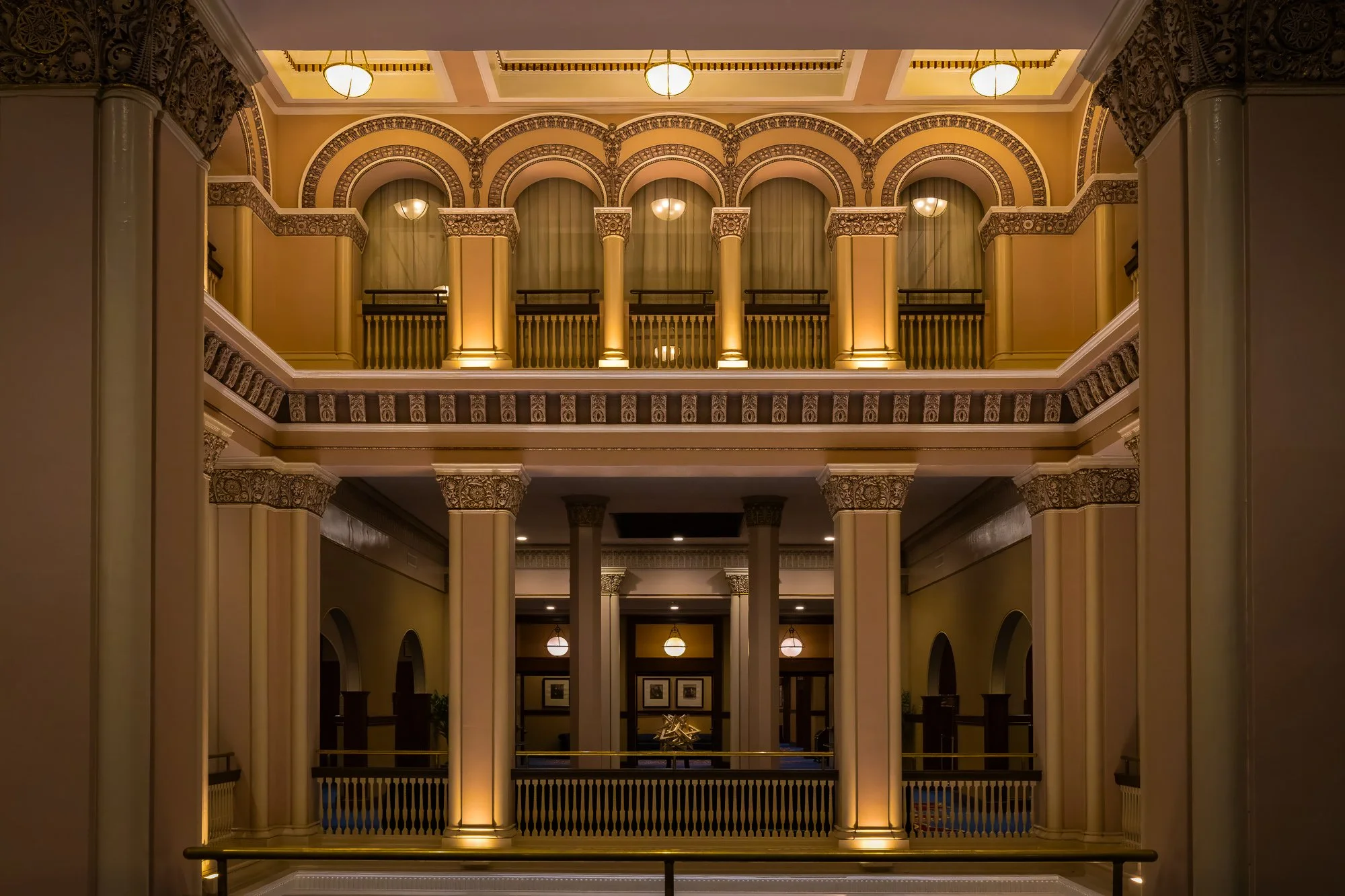 A symmetrical interior view of St. Louis Union Station featuring warm yellow lighting, multiple levels with arched walkways, decorative Romanesque columns, and intricate plasterwork along the ceilings and balconies.
