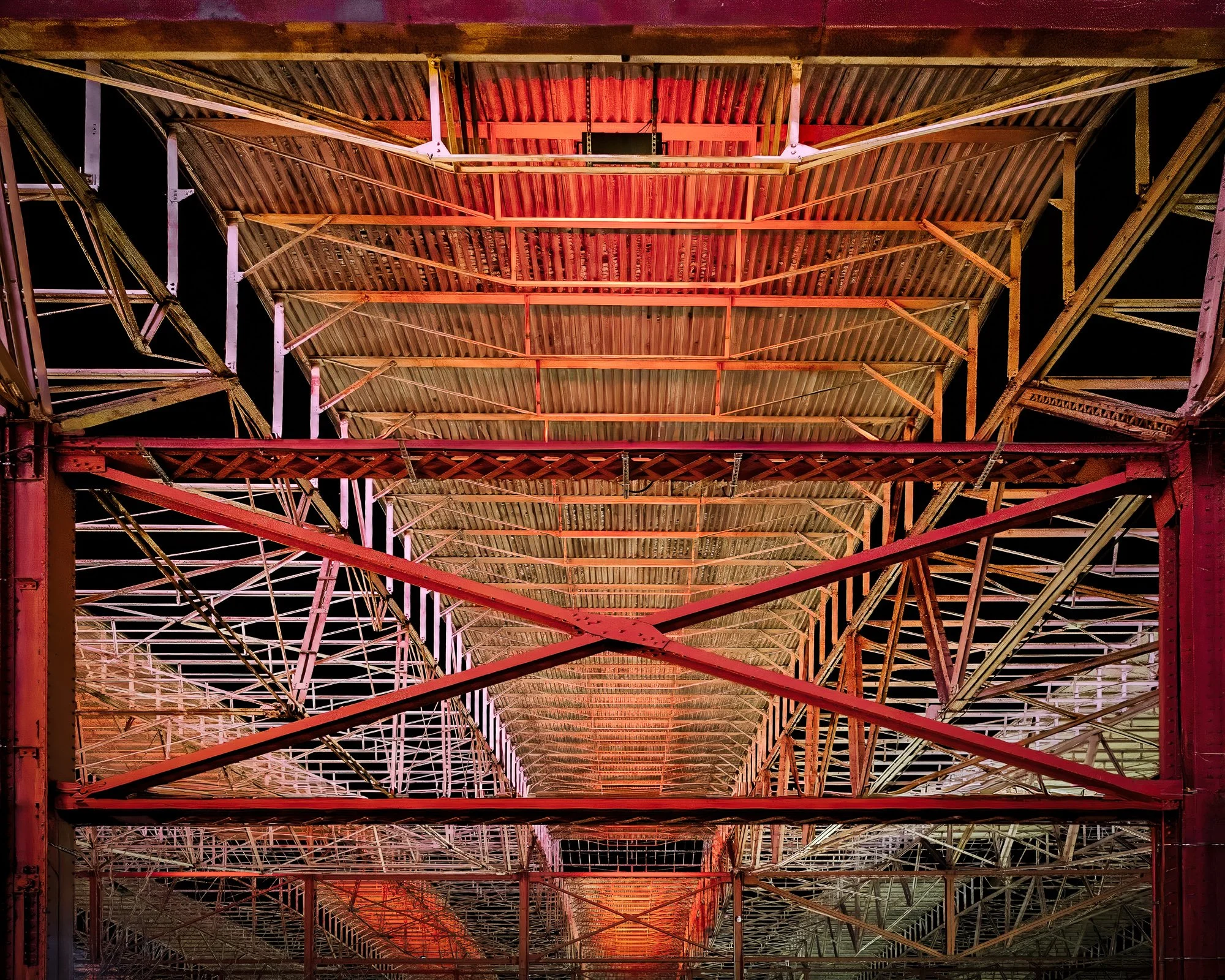 A photograph looking straight up at the underside of the St. Louis Union Station train shed. The complex network of red and orange illuminated steel and iron trusses forms a massive geometric pattern against the dark night sky.