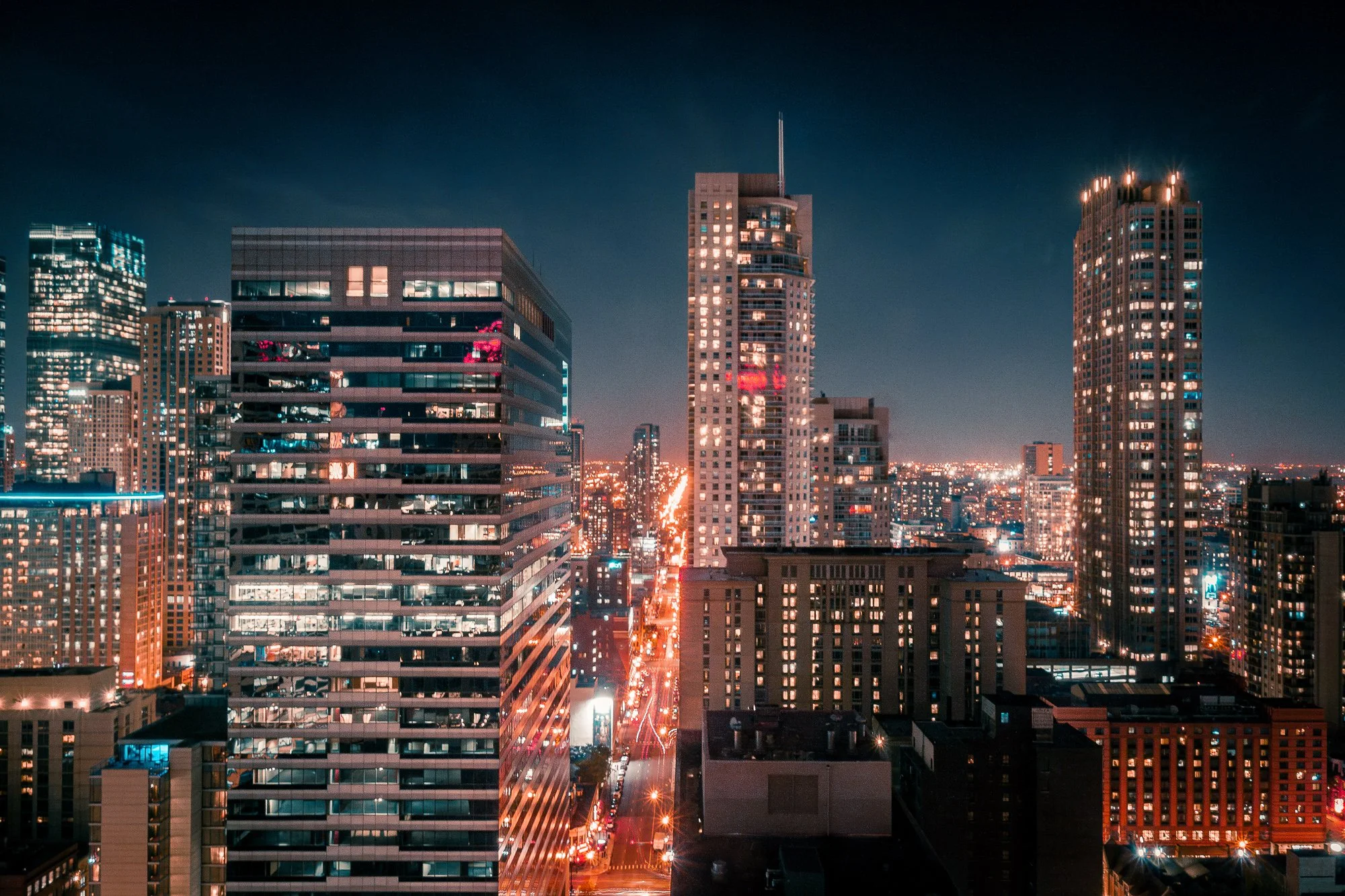 A vibrant photo of the Chicago skyline captures the glowing city streets lined with towering buildings. Warm orange streetlights contrast with the cool, bluish reflections on glass, creating a mesmerizing mix of modern architecture and urban energy.