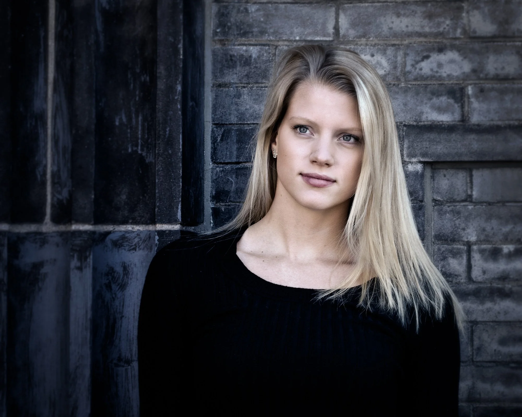 The image shows a woman with blonde hair standing against a textured brick wall. She has a calm expression, wearing a black top, with her hair cascading over her shoulder. Natural light highlights her features, creating a moody atmosphere.