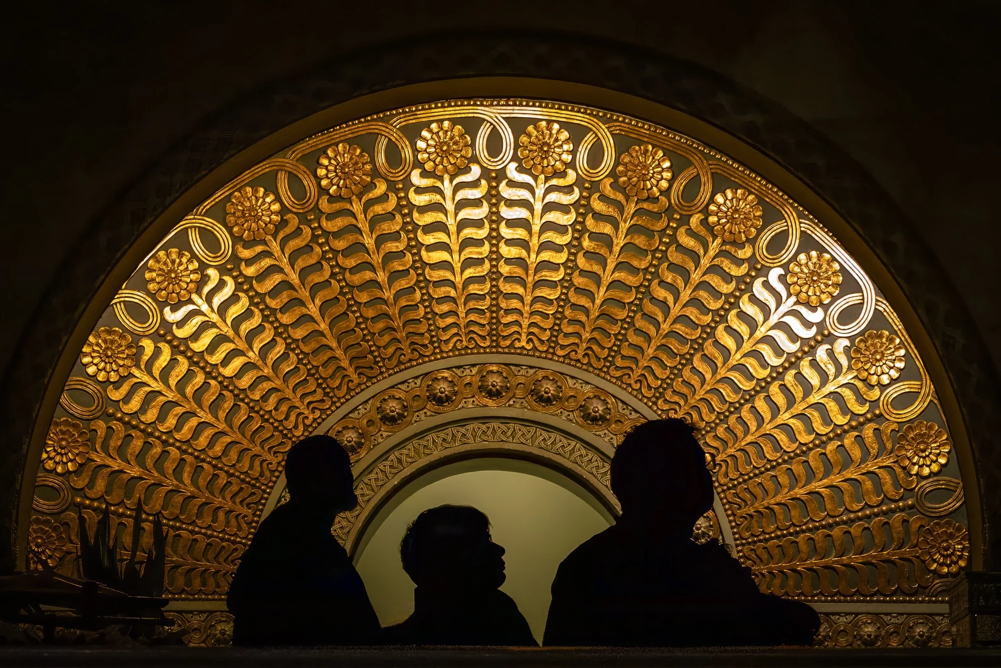 A photograph of a large, ornate semi-circular architectural detail covered in gold leaf floral patterns. The dark silhouettes of three people are visible in the foreground, contrasting against the brightly lit gold background.