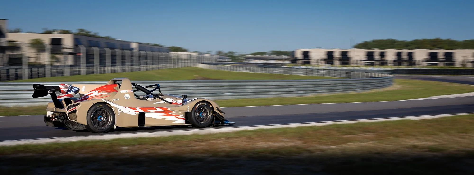 A beige and red Radical race car speeds down the track at The Motor Enclave in Tampa, Florida, captured in a wide panning shot that blurs the background.