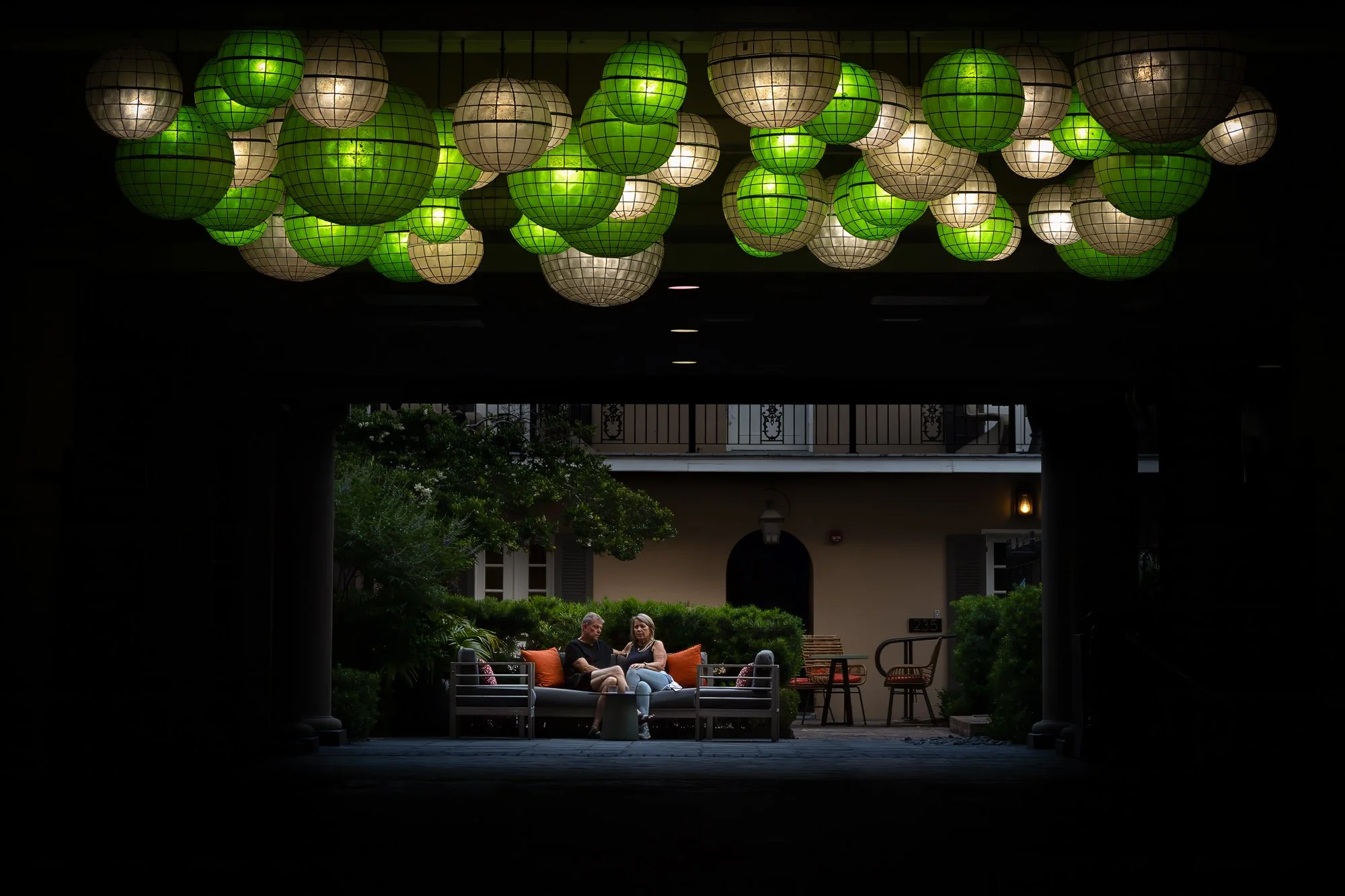 Green and white lanterns hang above a shadowed courtyard at Hotel de la Poste in the French Quarter, with two people seated in the distance.