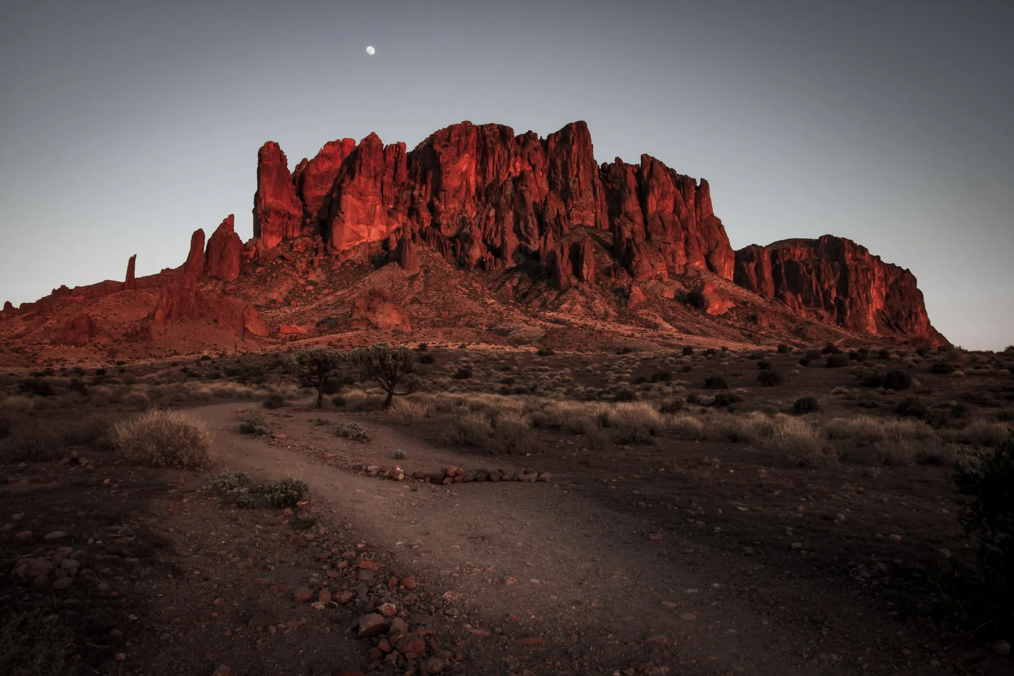The Superstition Mountains are bathed in warm, red light during sunset. The rugged peaks rise dramatically against a clear sky. The foreground features a winding dirt path bordered by desert vegetation and rocky terrain.