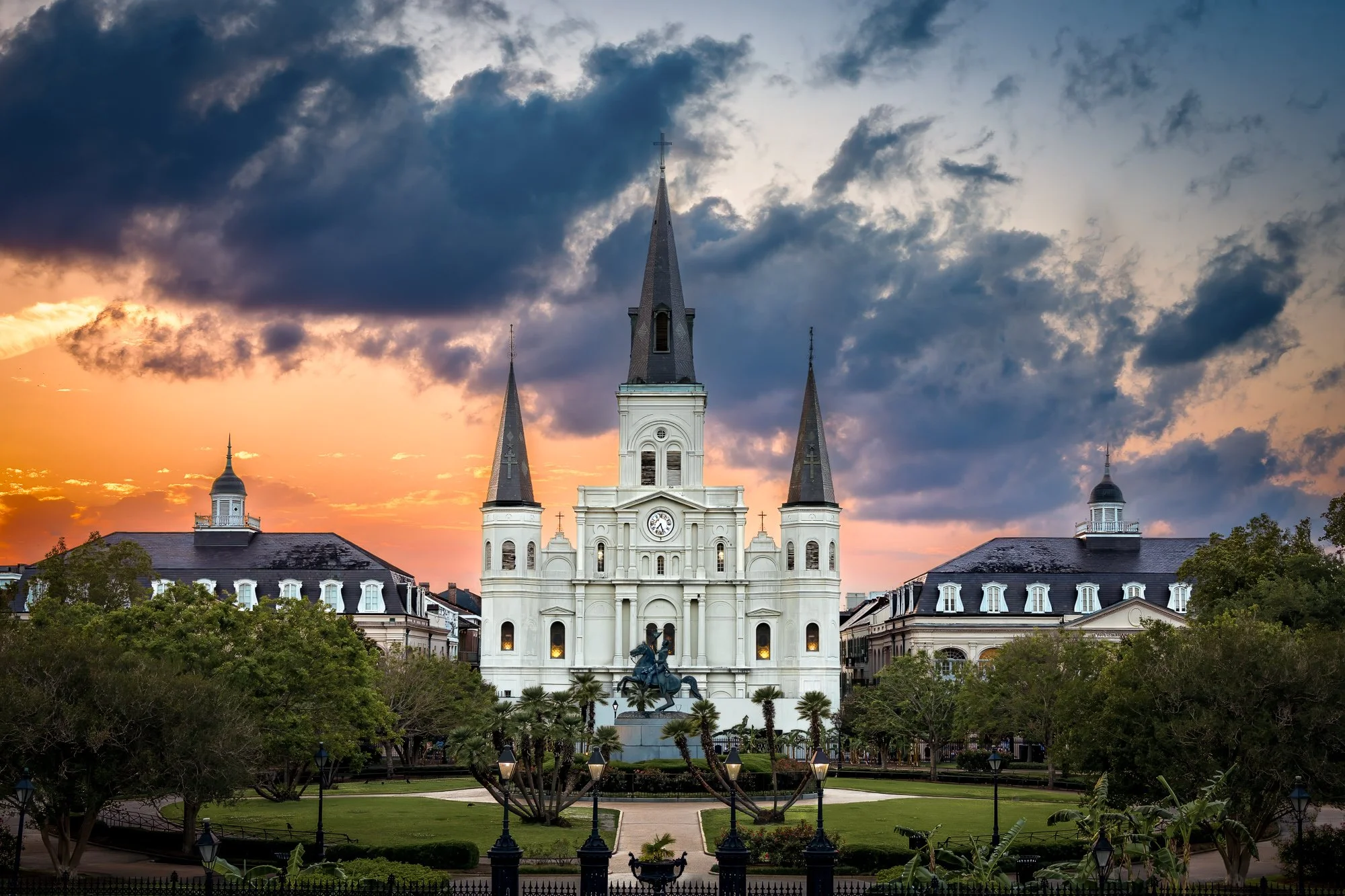 St. Louis Cathedral rises above Jackson Square in New Orleans at sunset, framed by dark clouds and warm evening color.