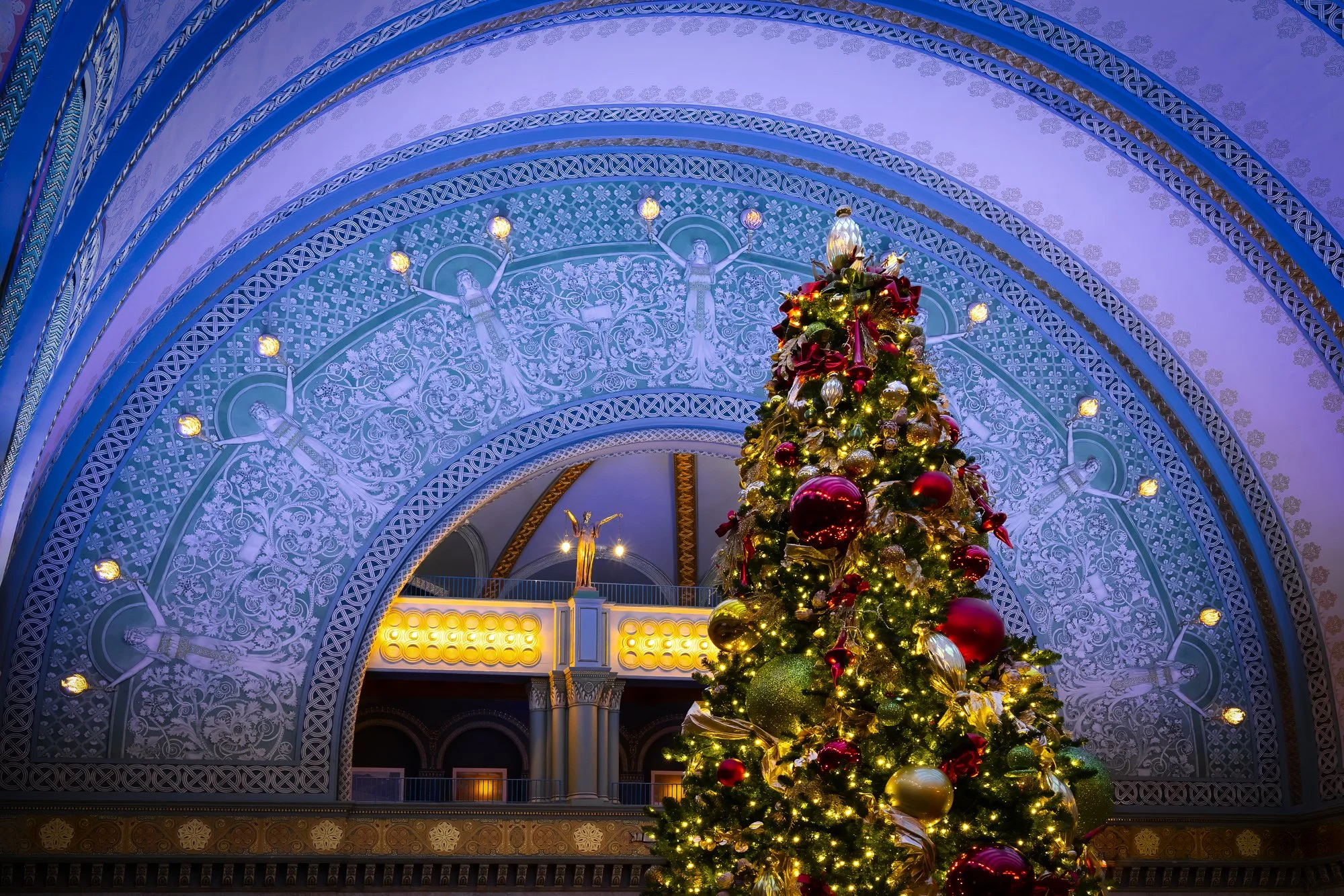 A large, brightly lit Christmas tree decorated with red and gold ornaments stands in the Grand Hall of St. Louis Union Station. The historic barrel-vaulted ceiling with intricate patterns and a stained glass window is visible in the background.
