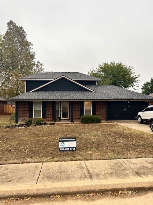 Two-story brick house with a new roof, featuring a sign from Elevated Roofing on the front lawn, a driveway with a parked car, and surrounding trees.