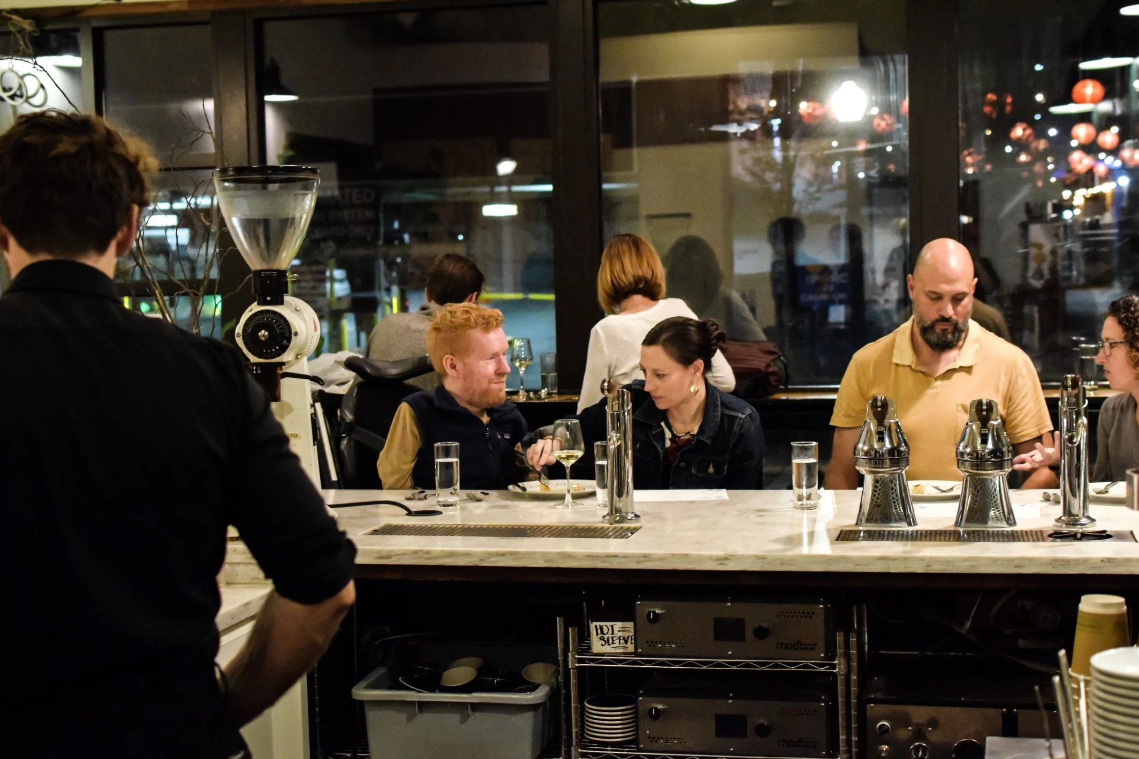 People dining at a restaurant bar counter with a bartender preparing drinks and others engaged in conversations.