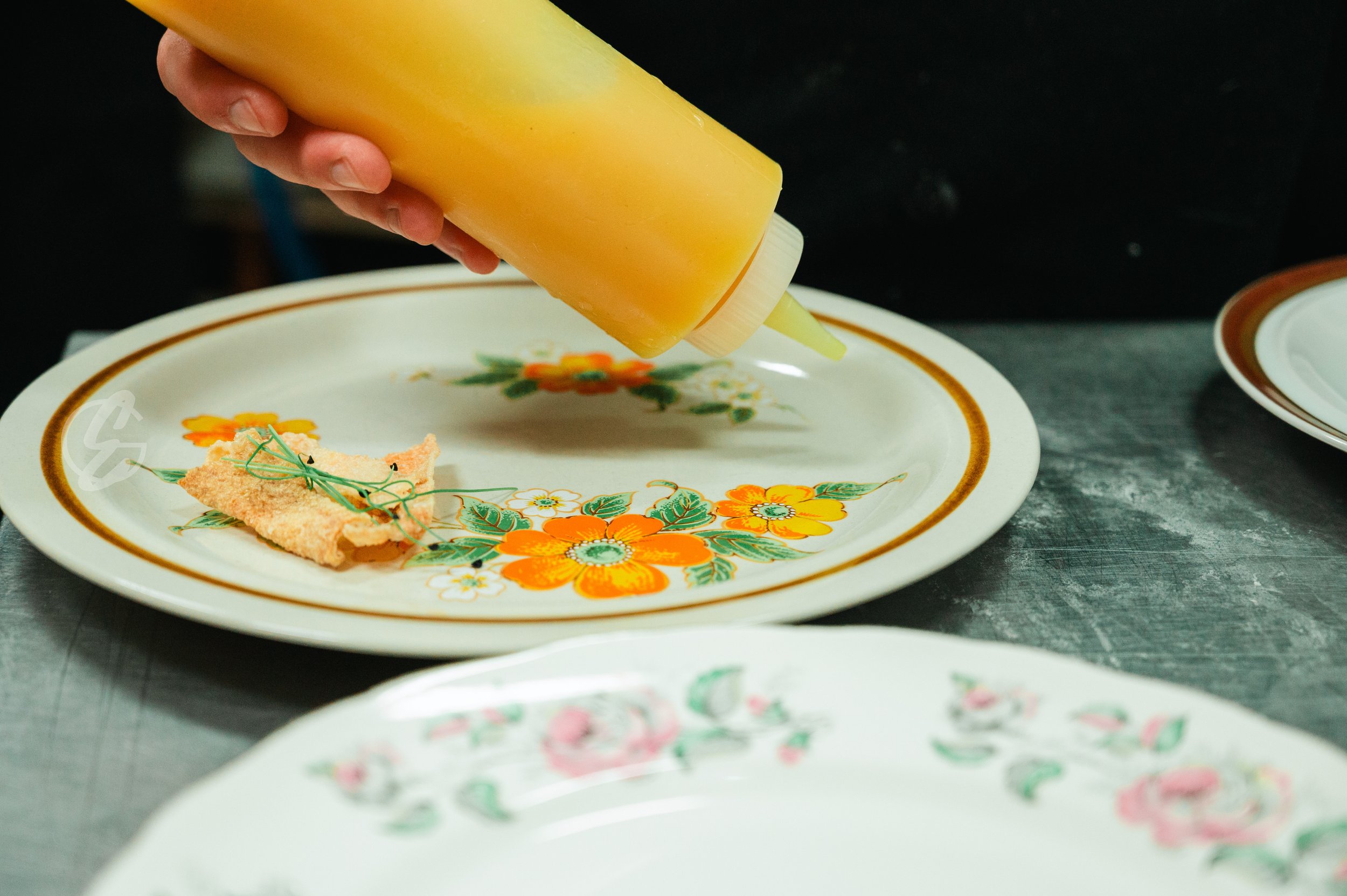 Person squeezing yellow mustard onto a floral patterned plate with a breaded food item and green garnish.