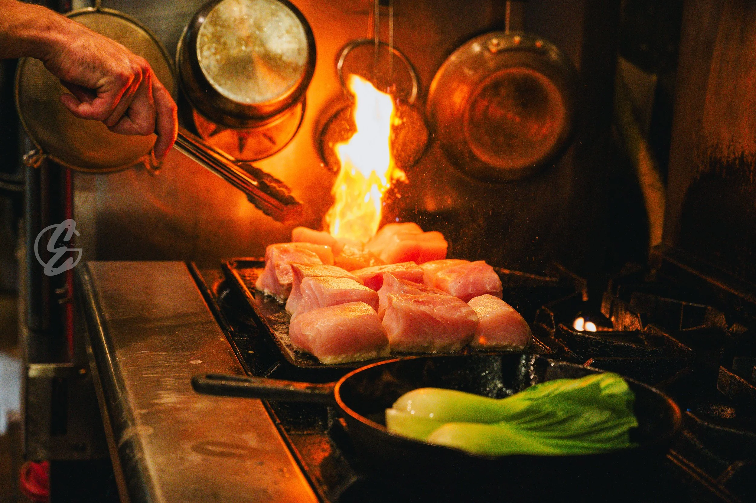 Cooking meat in a grill with a flame. A person is using tongs to handle the meat, with leafy greens in a pan nearby.