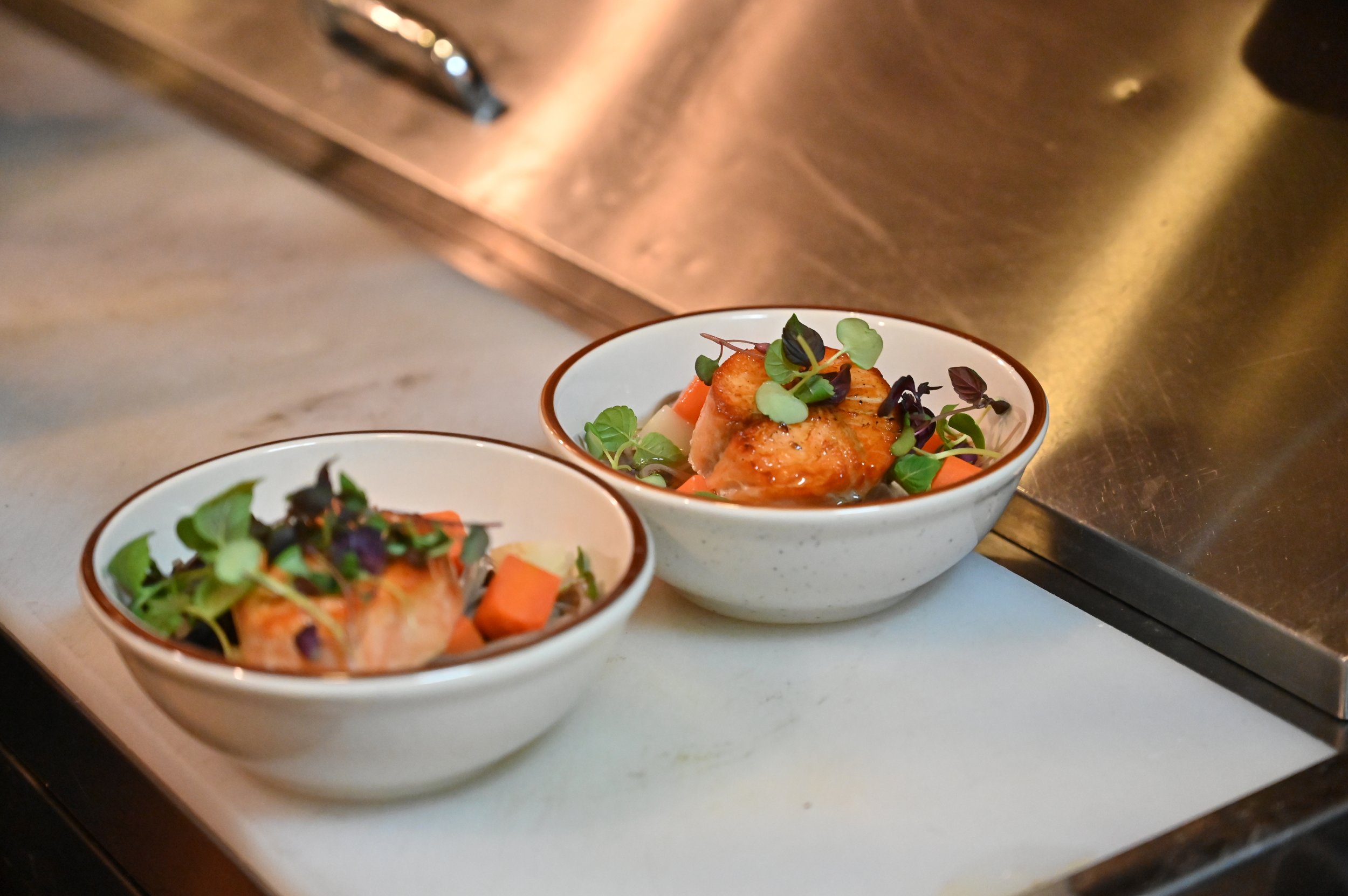 Two white bowls with grilled chicken, diced carrots, and microgreens on a white surface near a metal countertop.